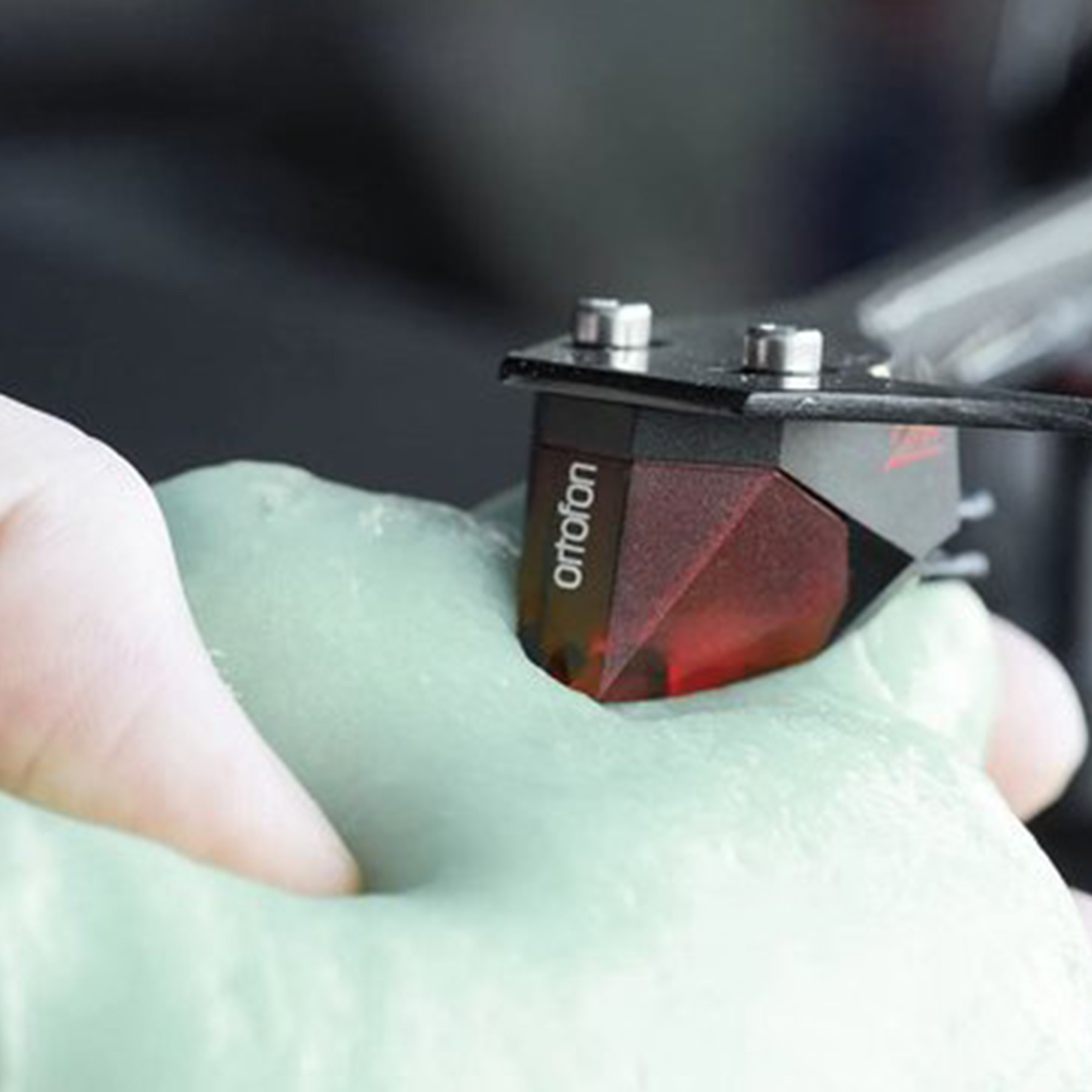 Dłoń trzyma wkład gramofonowy z czerwoną igłą, w kontakcie z zieloną substancją.