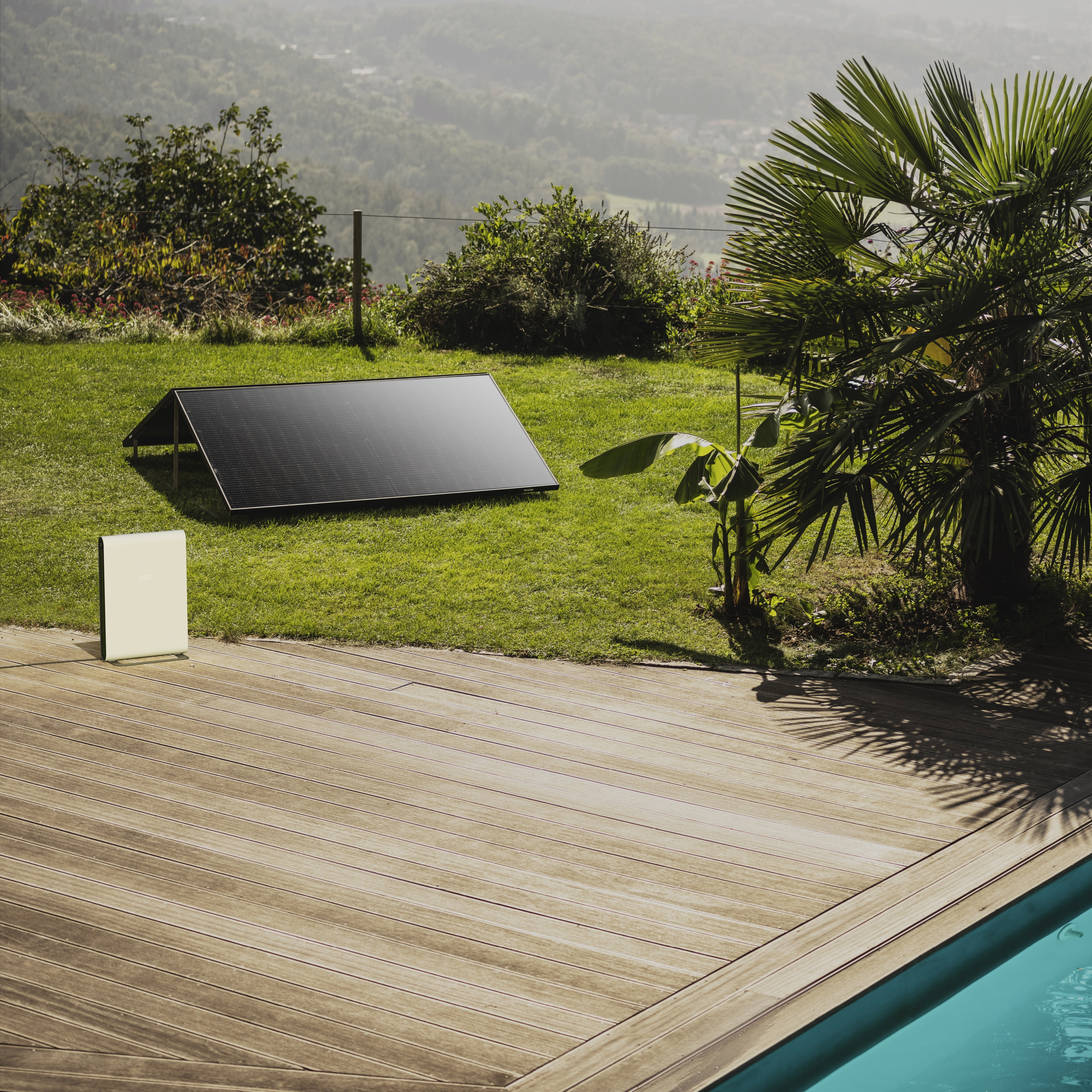 Solarpanel auf Gras, in der Nähe eines Holzdecks und Pools. Palmen und Berge im Hintergrund.