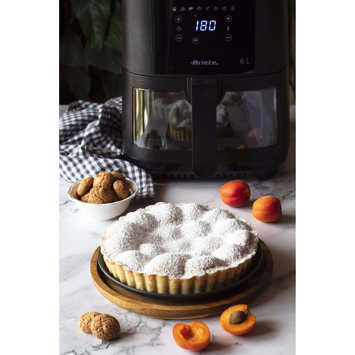 Frytownica powietrzna z ciastem, ciasteczkami i morelami na marmurowej powierzchni obok kraciastej szmatki.