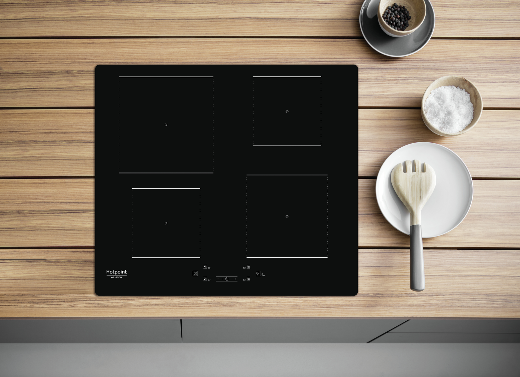 Czarna płyta indukcyjna z czterema palnikami, drewniana łyżka, sól i pieprz z boku.
