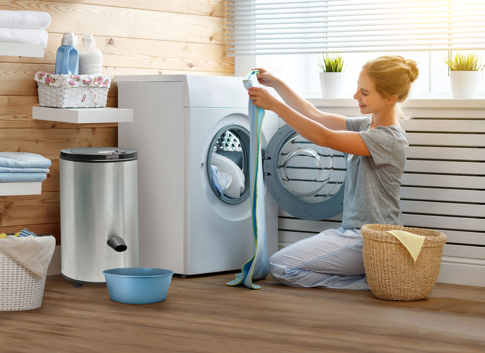 A woman puts a towel into a washing machine. There is a basket, trash can, and washing supplies.