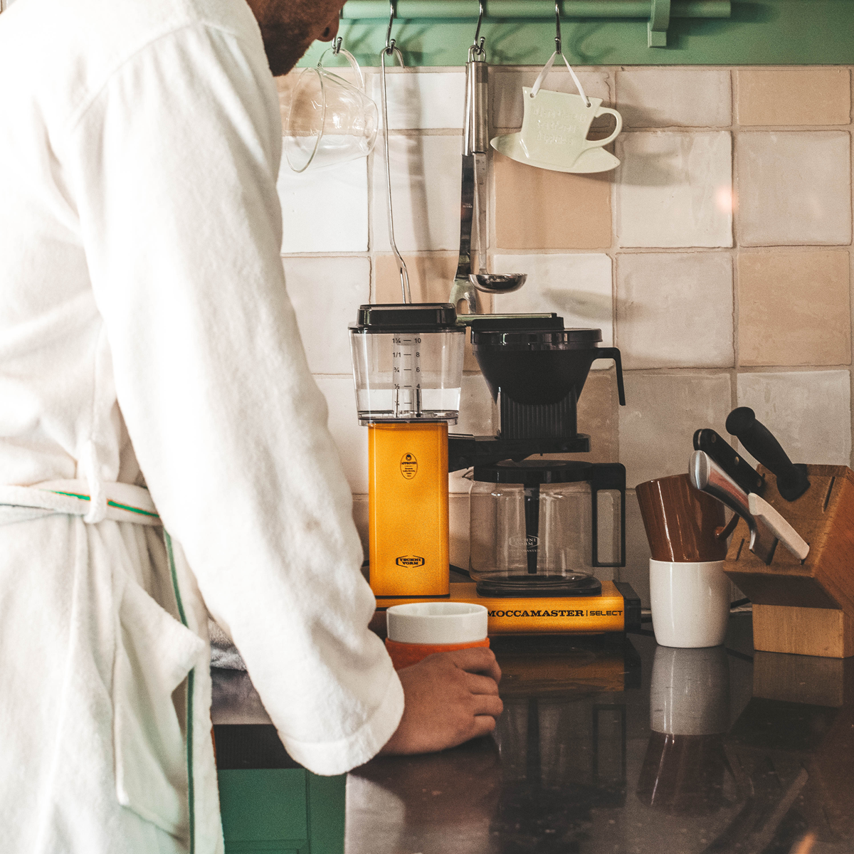 Mężczyzna w szlafroku robi kawę z ekspresu. Ekspres do kawy jest żółty.