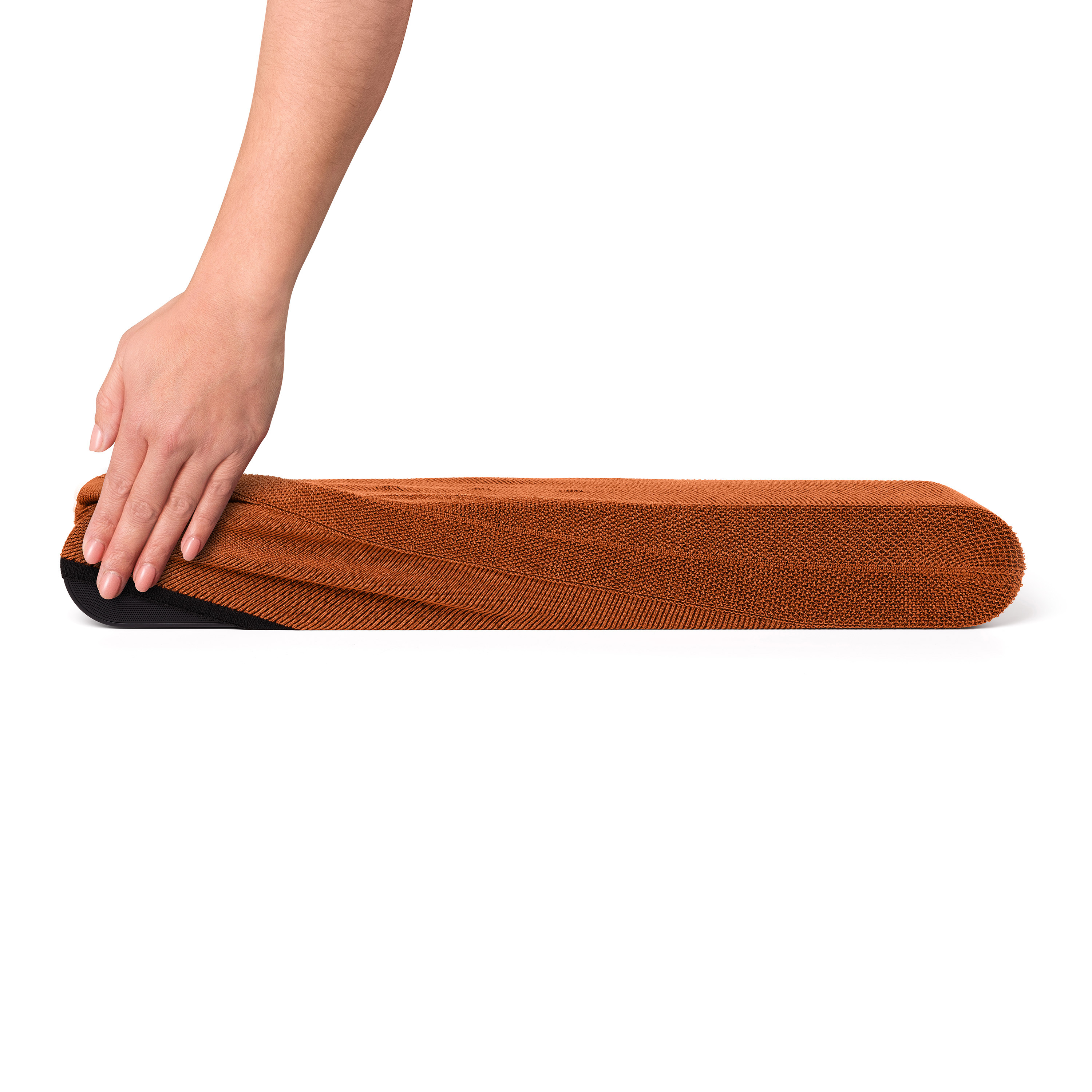 Main plaçant un tapis marron plié sur une surface blanche. Le tapis est orange. Dessous noir.