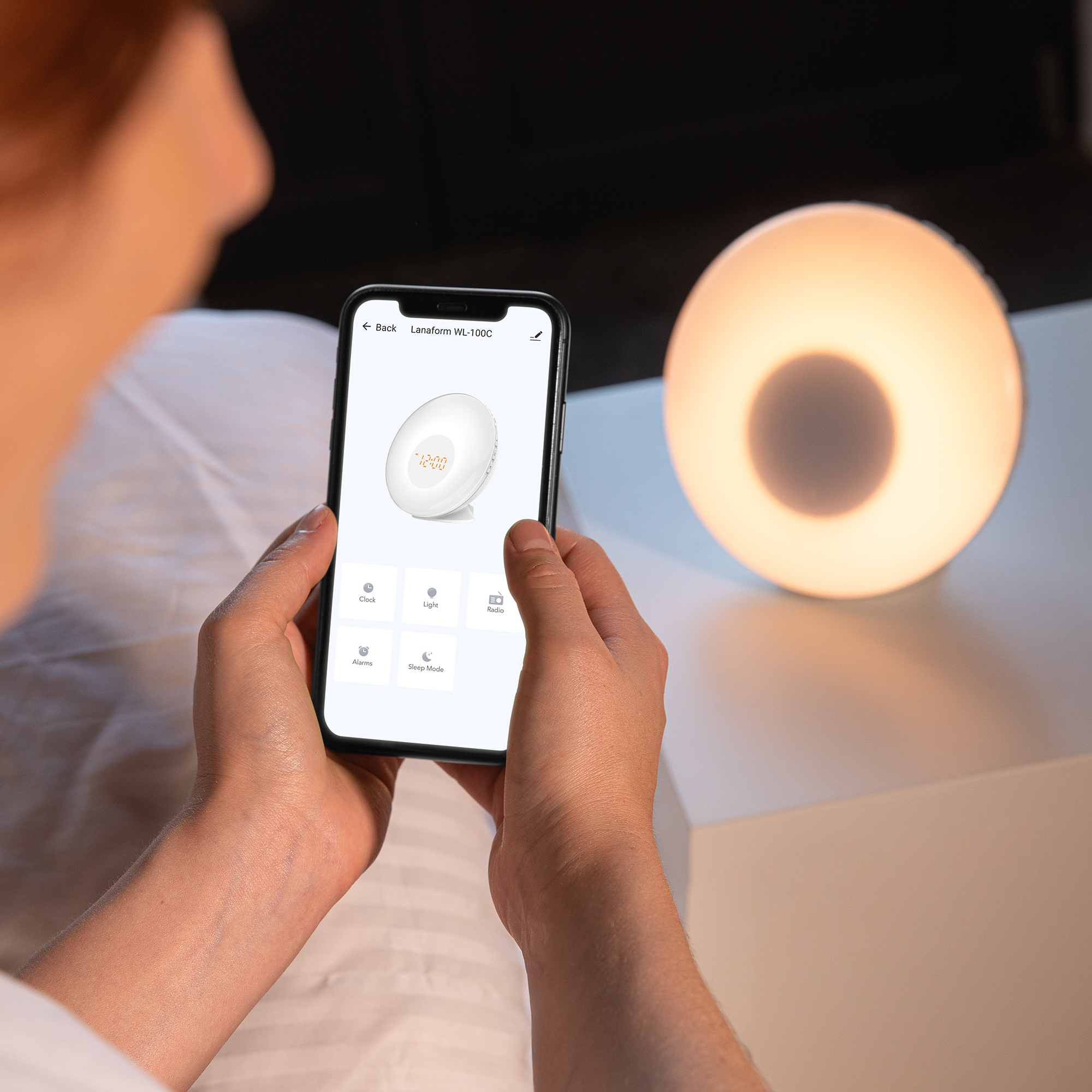 A person holding a smartphone displaying a smart alarm clock with a round light on a white surface.