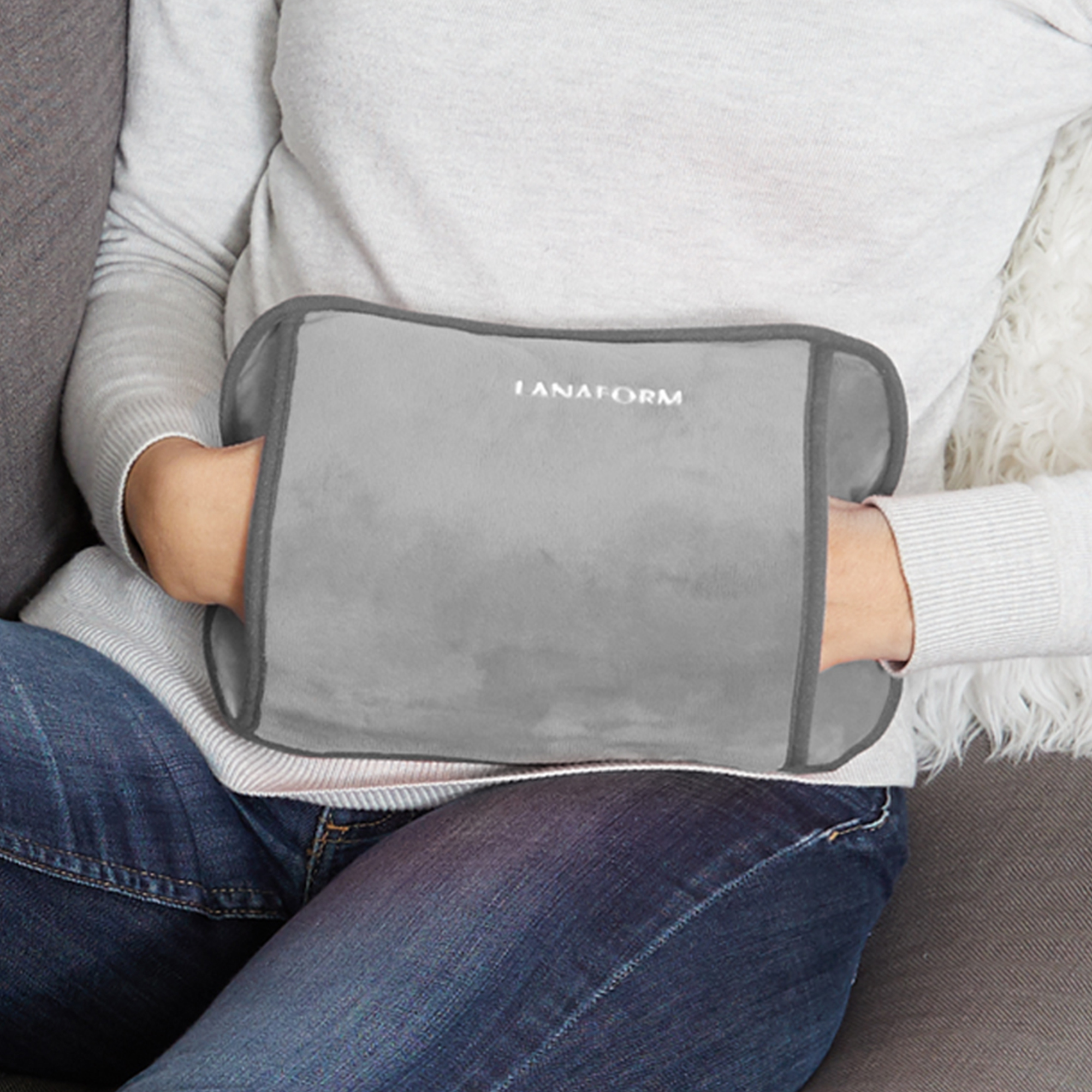 Woman holding a gray heating pad on her stomach, while sitting on a sofa.