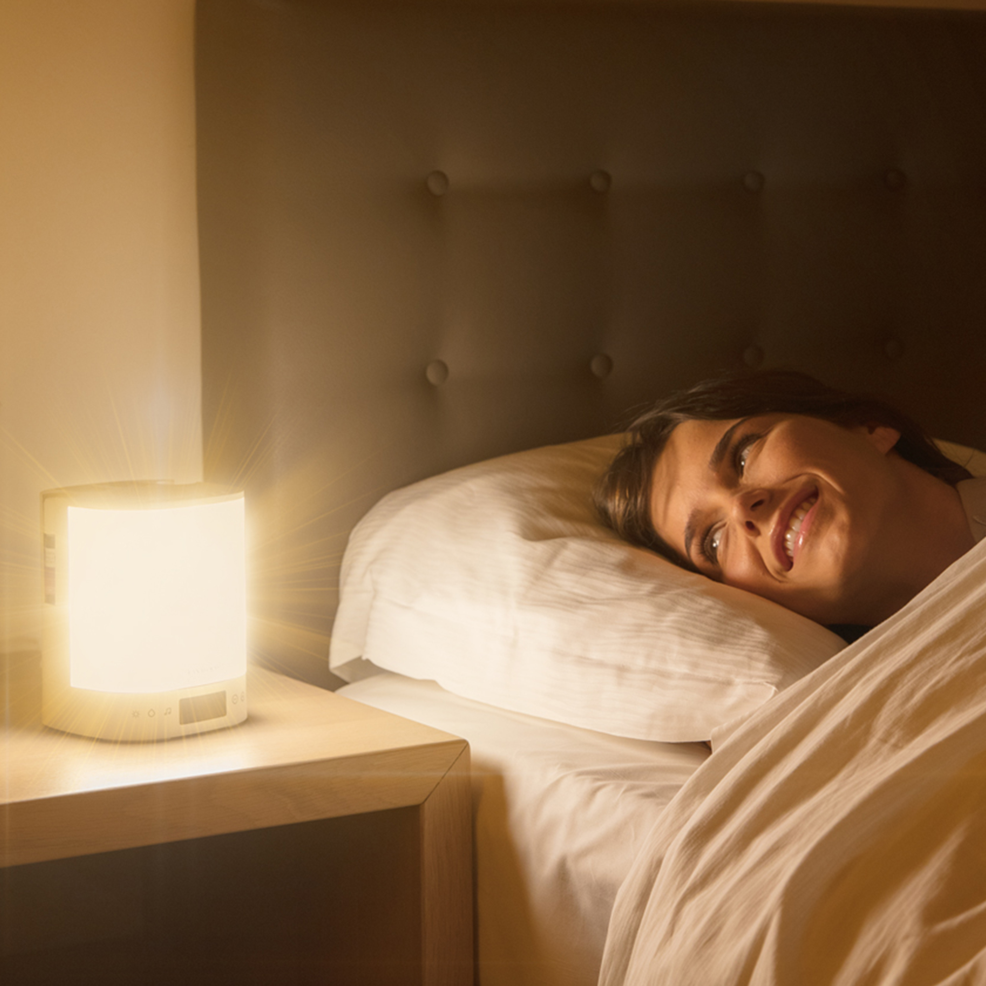 A woman in bed smiles at a bedside lamp. The lamp shines light. A white bed and a wooden table are in the shot.