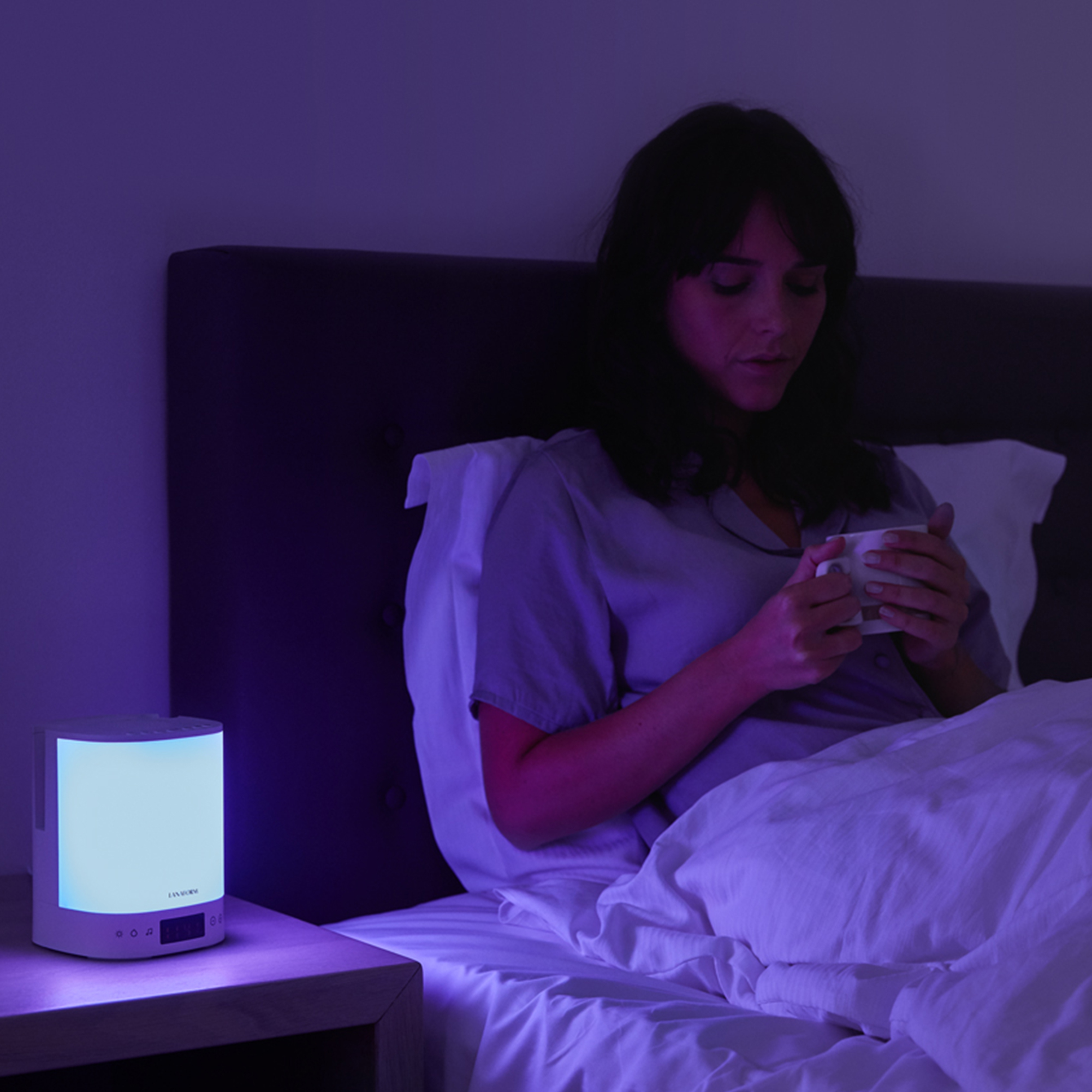 Woman in bed with a light-up bedside lamp. She is holding a cup and is in a dark room.