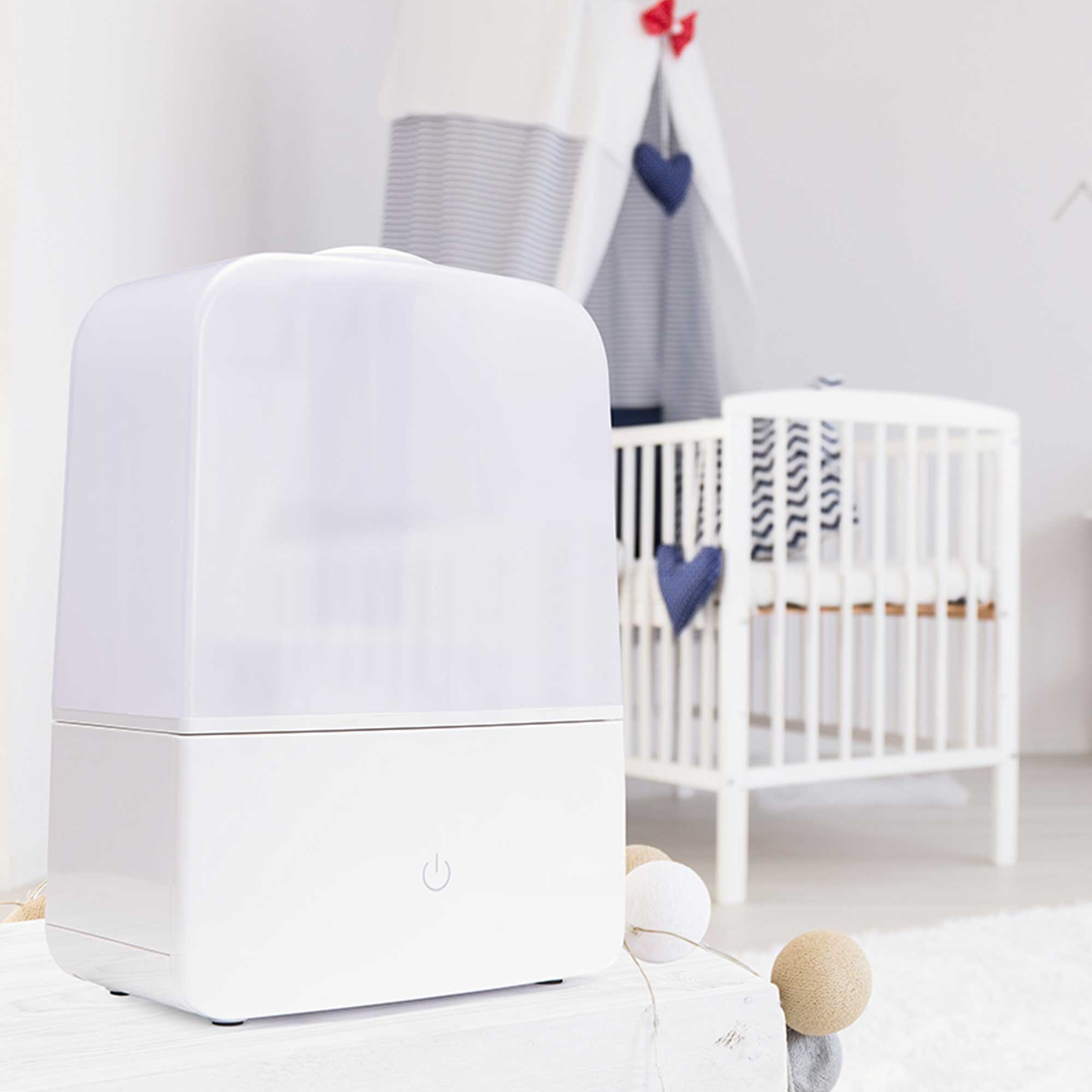 Weißer Luftbefeuchter auf einem Tisch. Im Hintergrund ein Kinderbett und ein Tipi. Die Szene ist in einem weißen Raum.