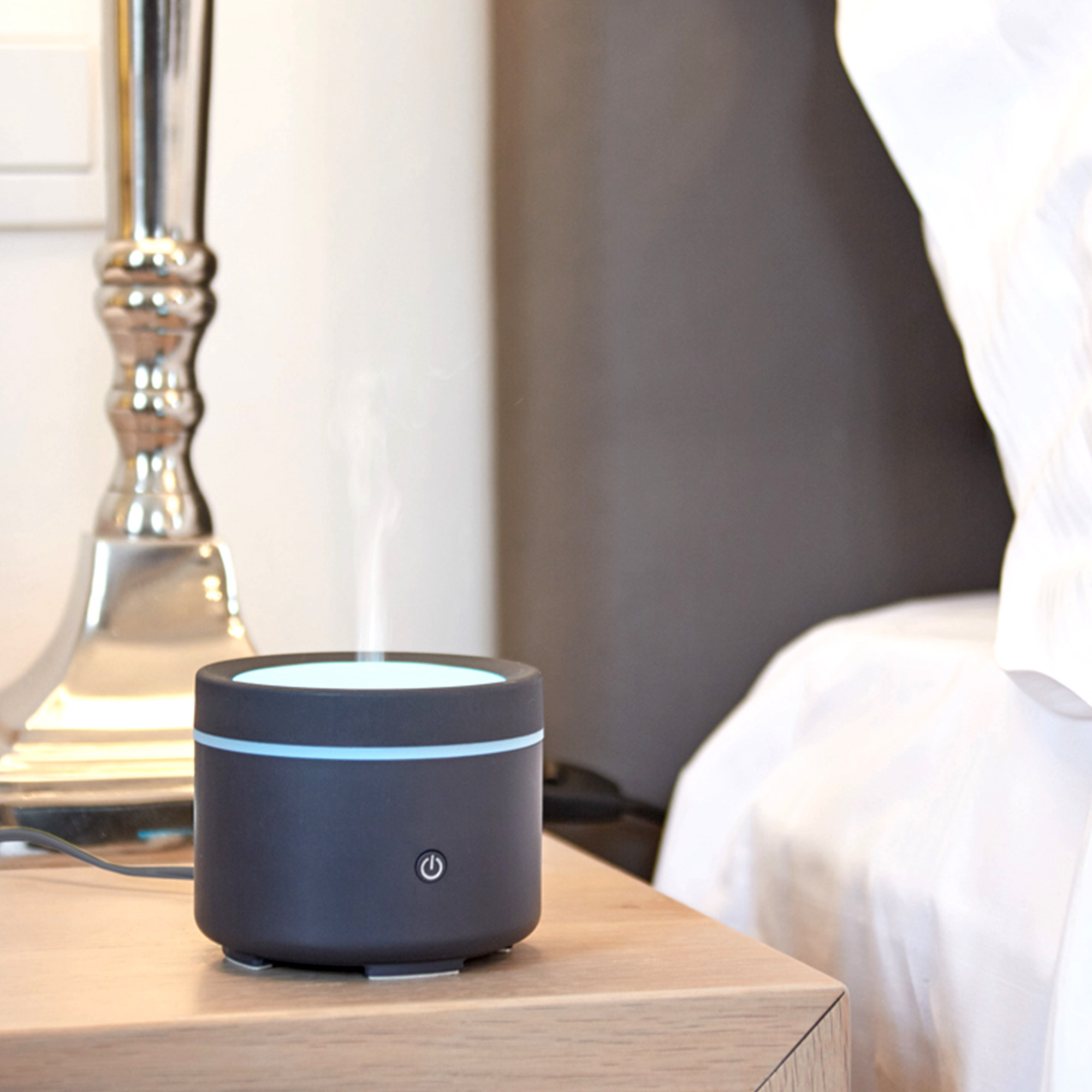 Black and gray aromatherapy diffuser on a wooden table, next to a lamp and a bed.