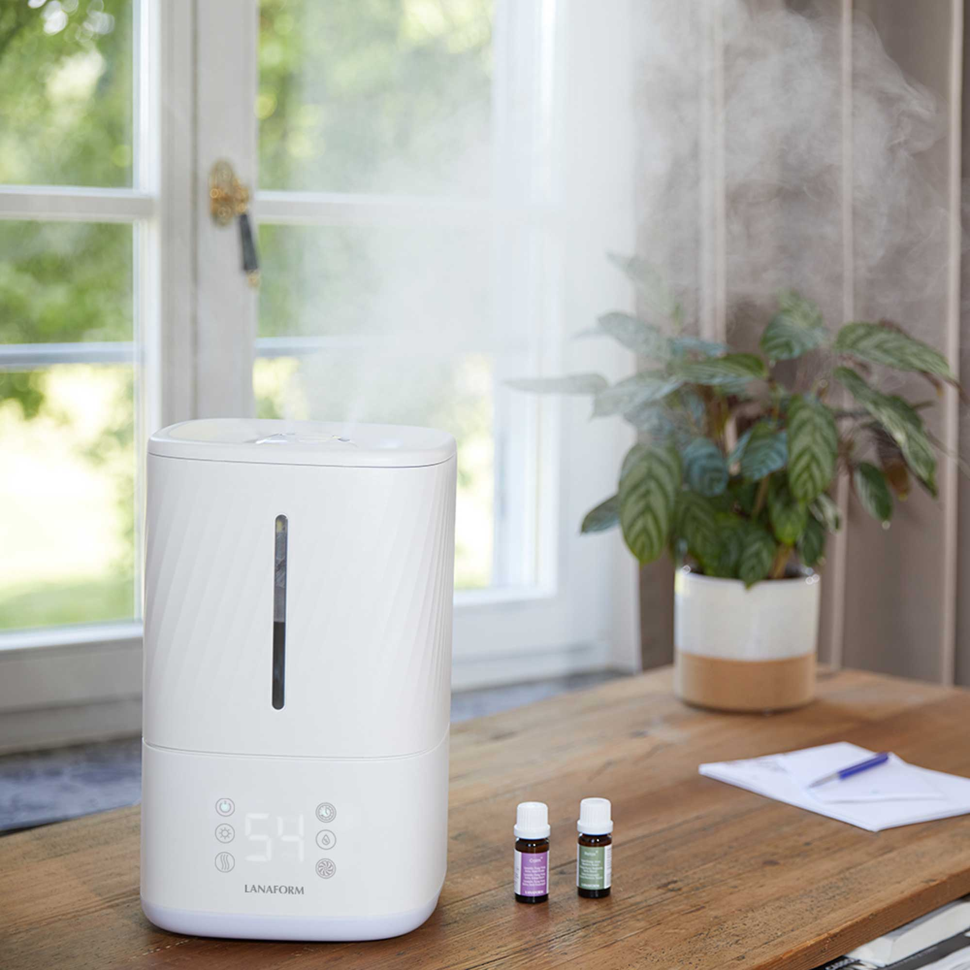 White humidifier on a wooden table. A plant, essential oils, and a pen and paper are nearby.