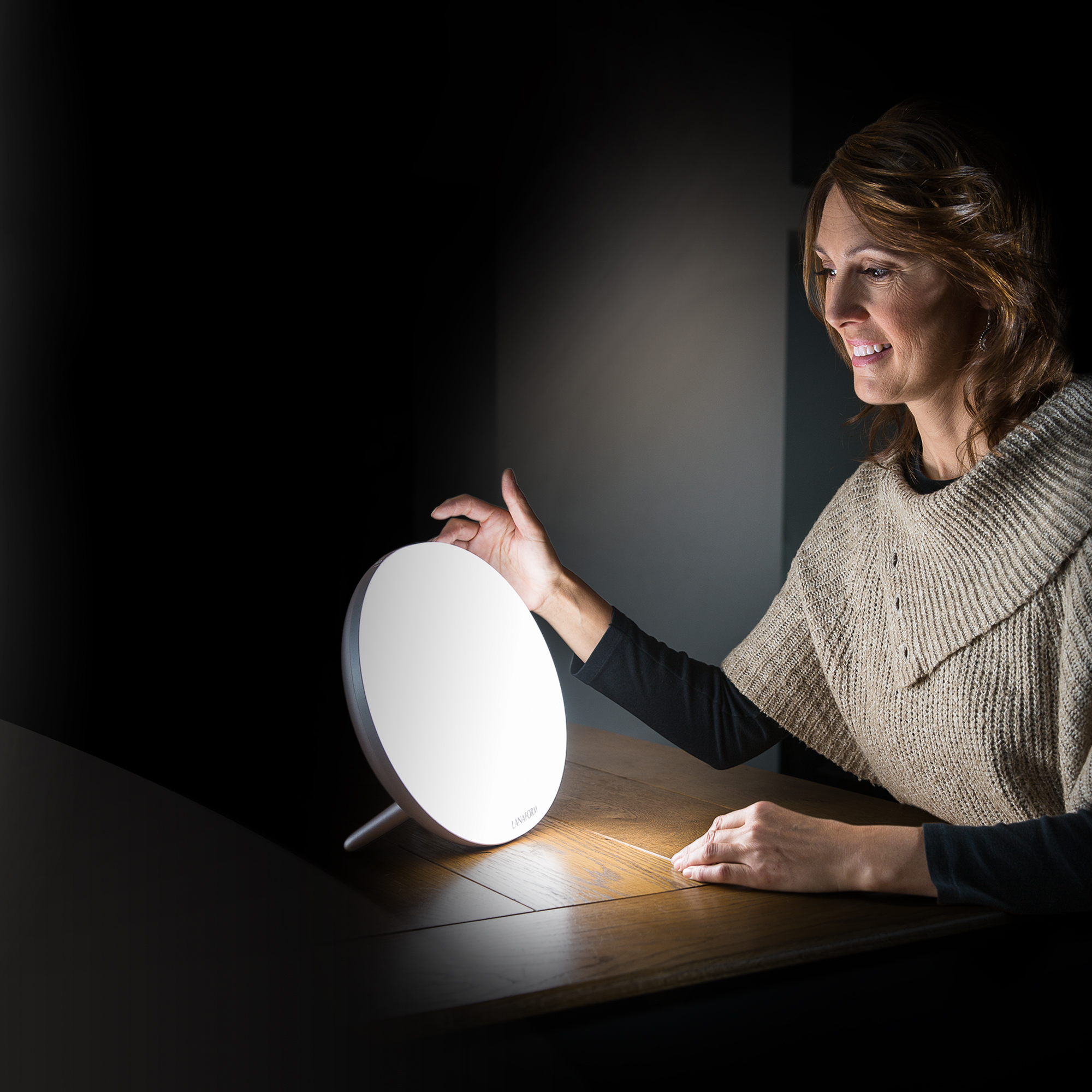A woman touches a bright white circular lamp, looking at it with a smile, in a dark room.