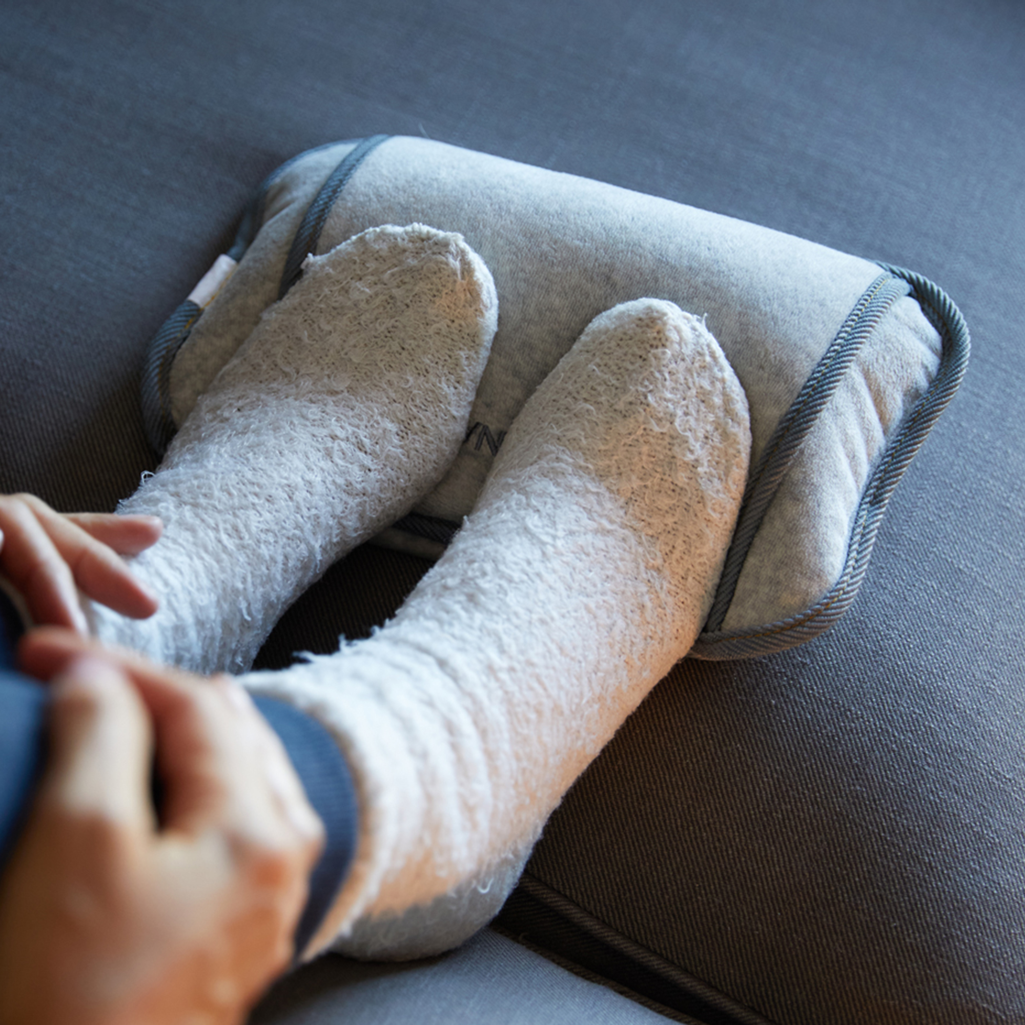 Füße in flauschigen weißen Socken auf einem grauen Kissen auf einer grauen Couch. Eine Hand ist sichtbar.