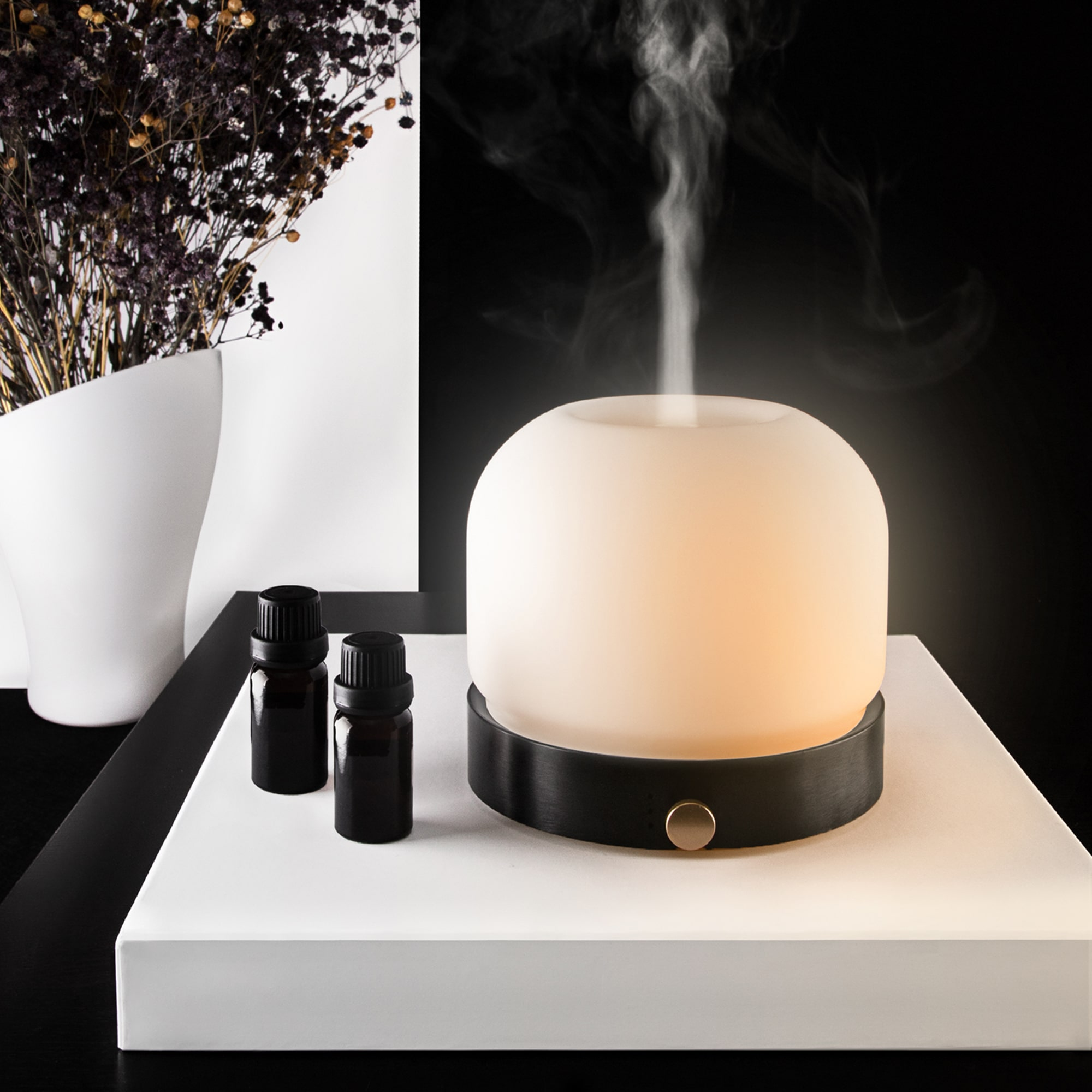 Aroma diffuser with vapor emitting. Two small bottles sit beside it. Black background and white surface.
