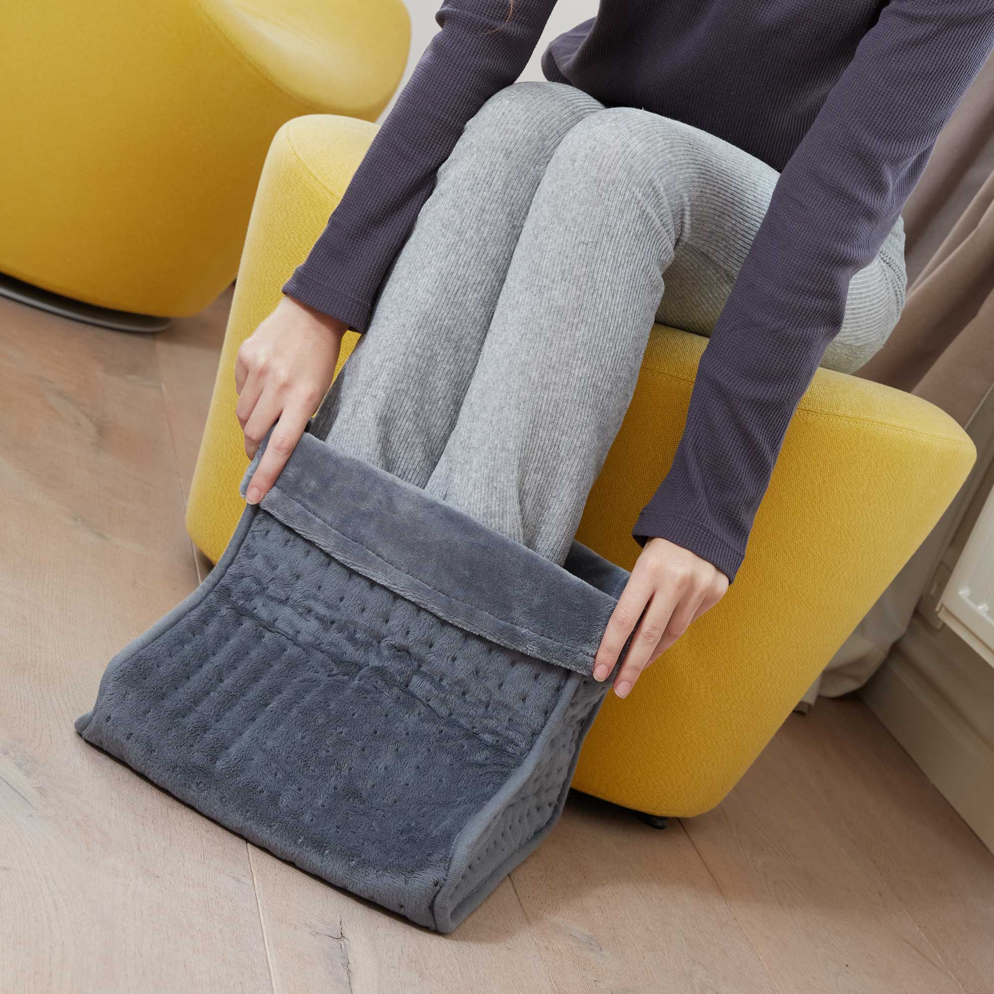 Osoba siedzi na żółtym krześle i zakłada na stopy szary podgrzewacz do stóp. Tło to neutralna ściana.