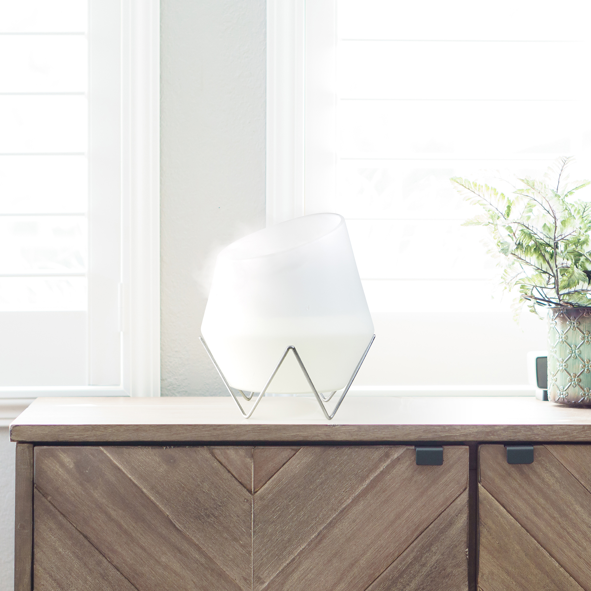 Biała szklana lampa z metalowym stojakiem na drewnianej szafce. Okno w tle.