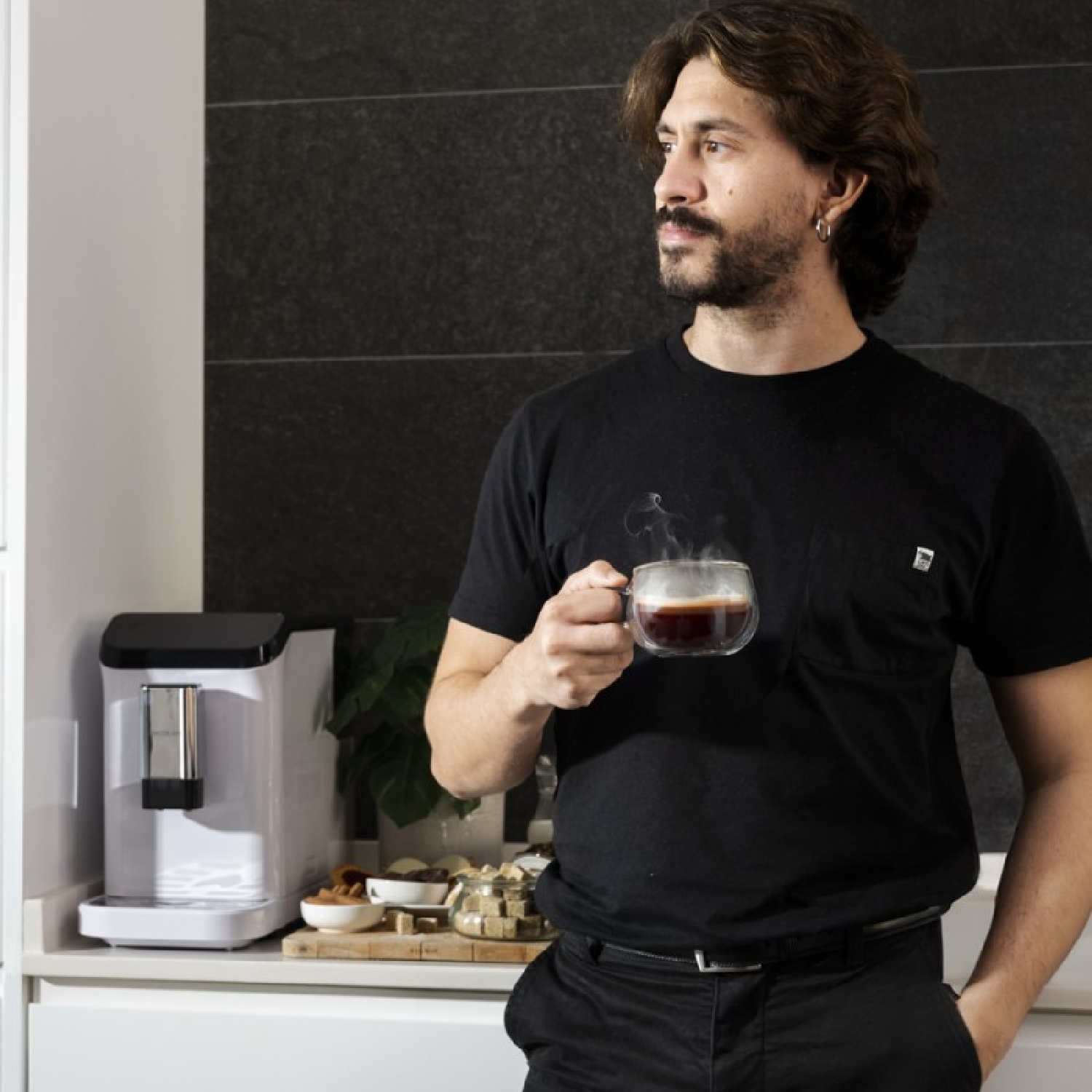 Hombre sosteniendo café, mirando hacia otro lado, cerca de una cafetera blanca. Cocina.