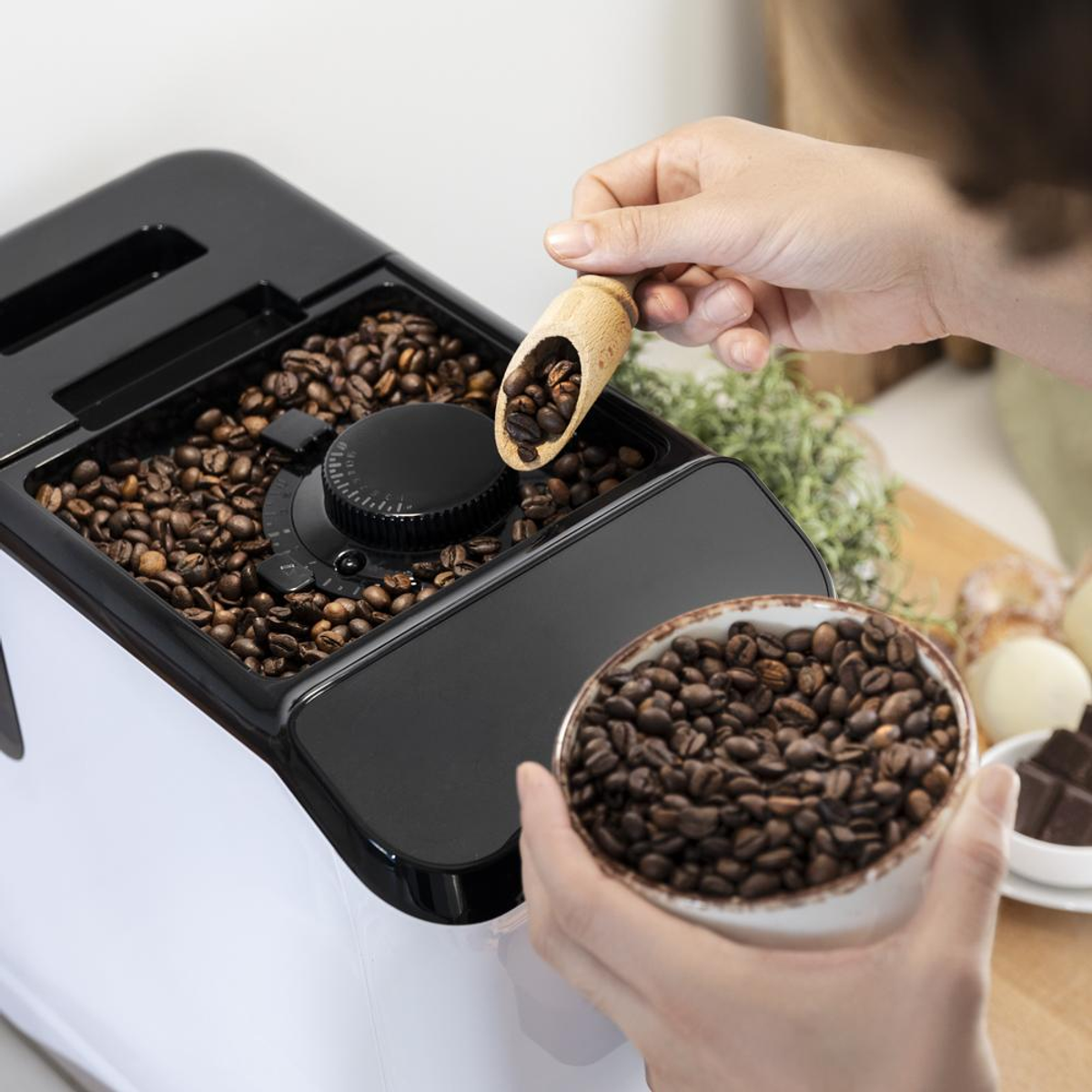 Persona usando un molinillo de café. El molinillo está lleno de granos de café. Cuchara de madera y un tazón de café.