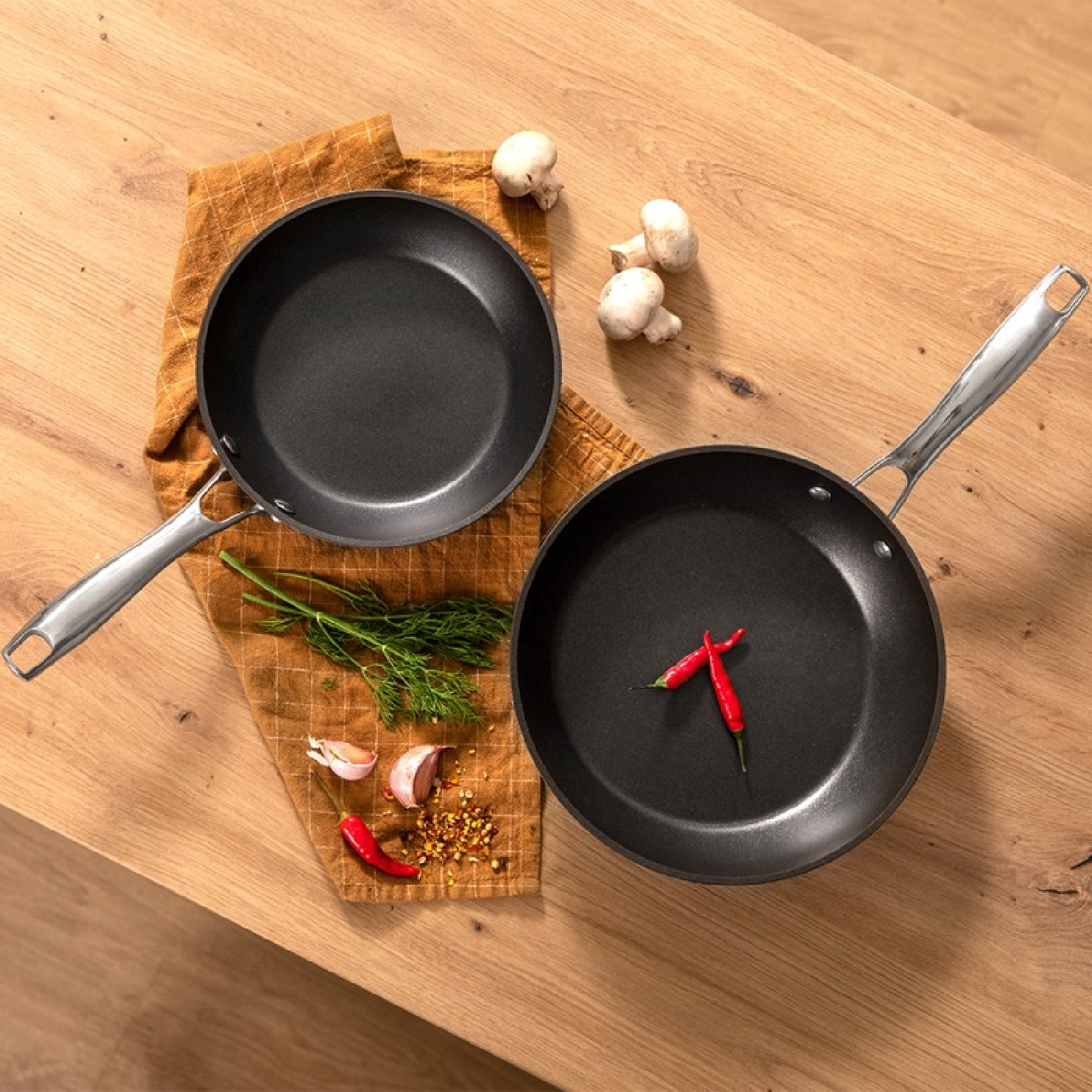 Twee zwarte koekenpannen met zilveren handvatten op een houten oppervlak. Ingrediënten zijn in de buurt.