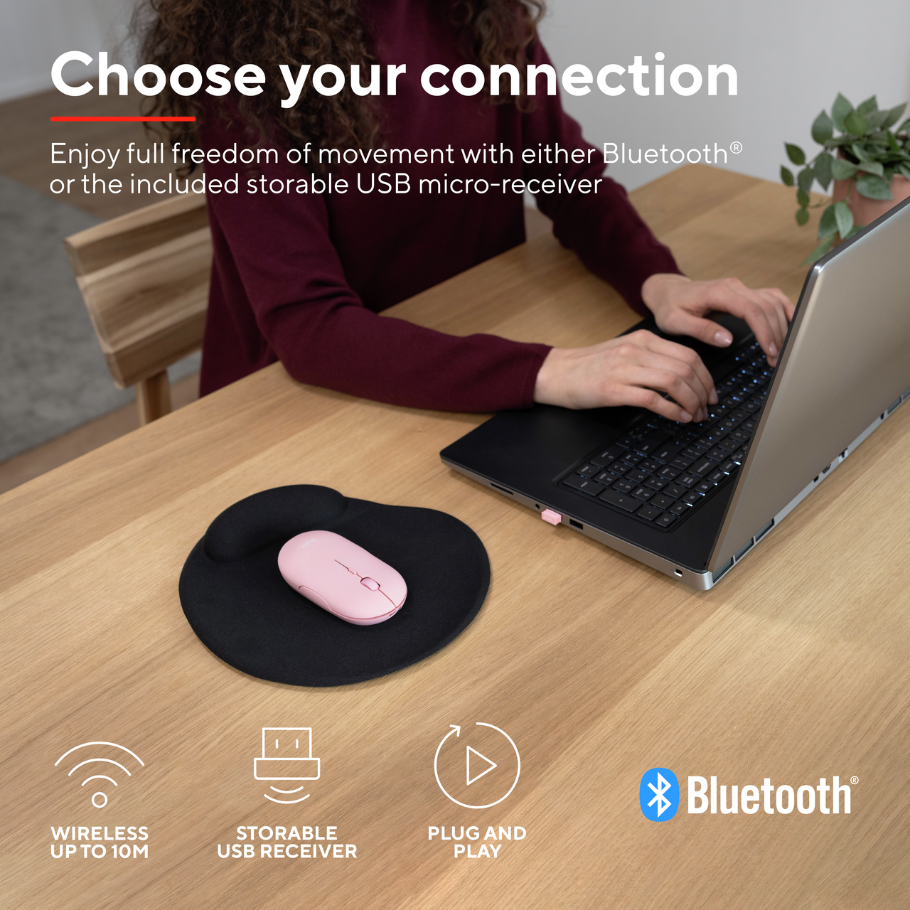 Persoon die een laptop gebruikt met een roze muis en een zwart muismat op een houten tafel.