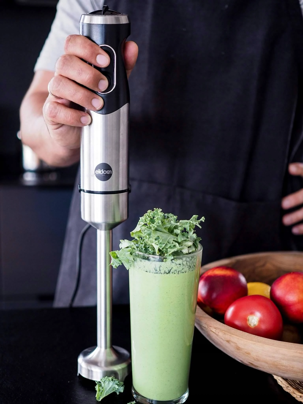 Een blender met groene smoothie, boerenkool en fruitschaal op een zwart oppervlak.