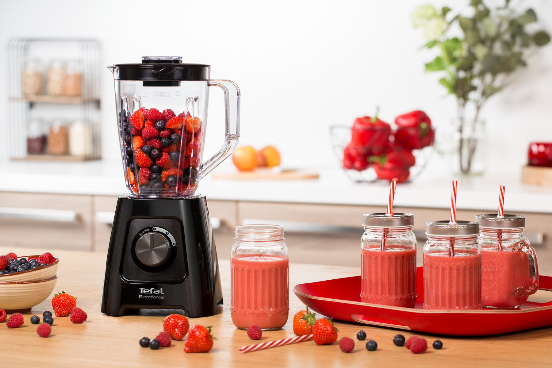 Licuadora con fruta dentro, tres batidos en frascos y fruta en una mesa de madera. Cocina luminosa.