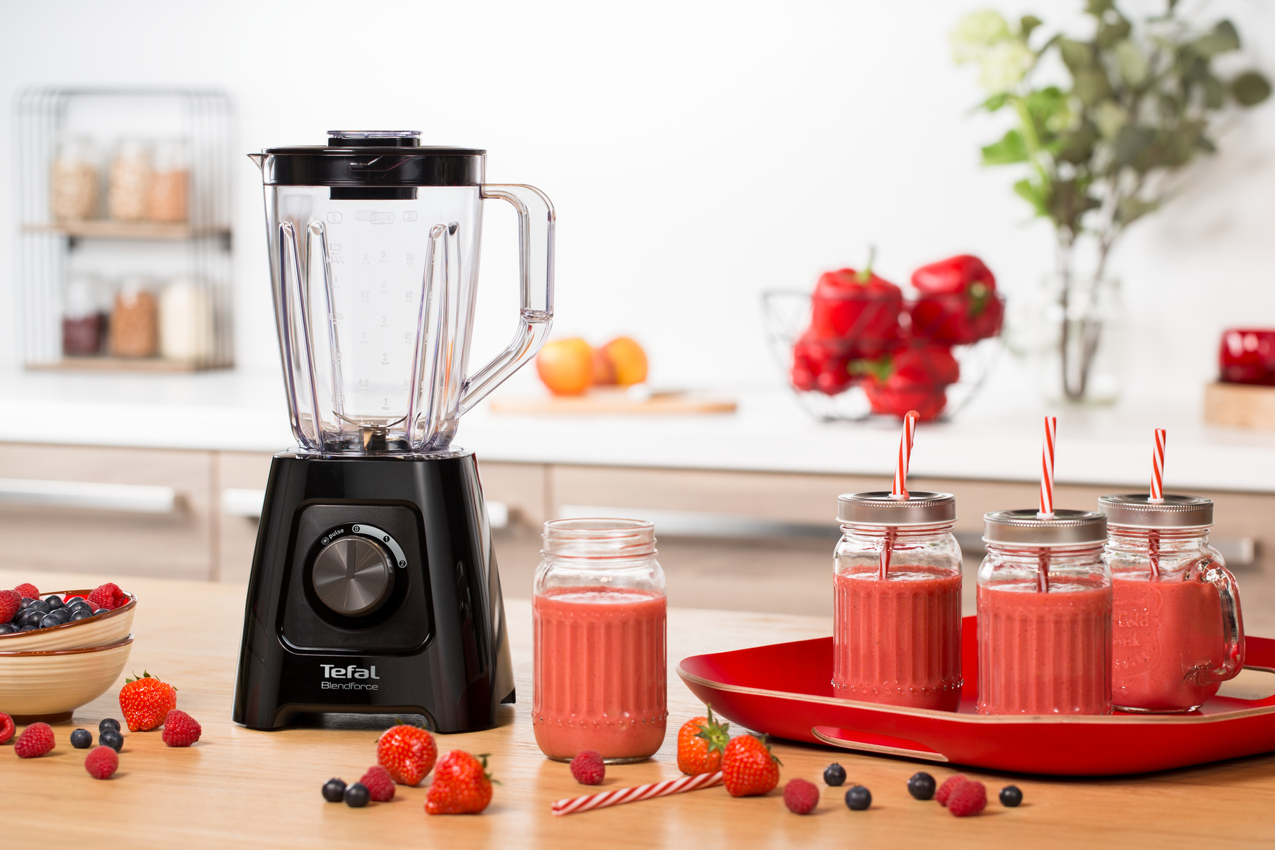 Licuadora con vasos de batido, fresas, arándanos y pimientos rojos en mesa de madera.
