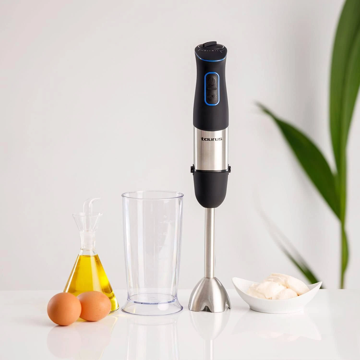 Batidora de mano con ingredientes: aceite, huevos y un cuenco de comida, sobre superficie blanca.
