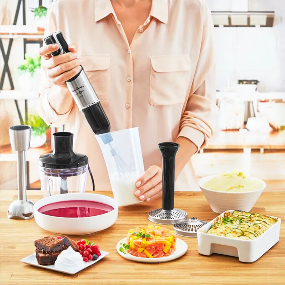 Una donna usa un frullatore con vari ingredienti sul bancone. Ci sono anche altri utensili da cucina.
