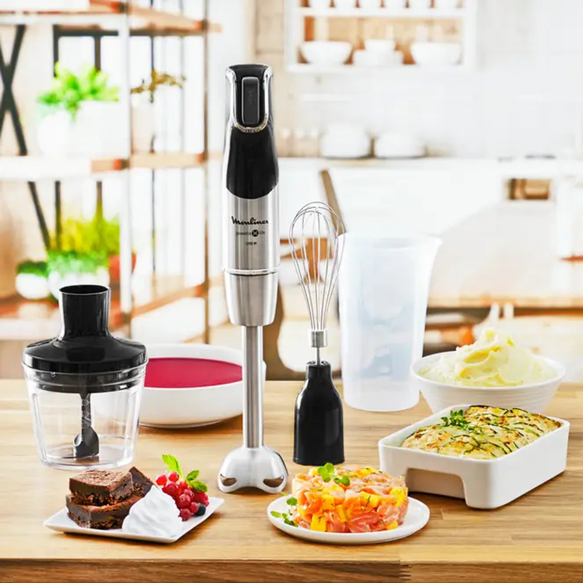 Frullatore a immersione con accessori su un tavolo vicino a cibo e sfondo cucina.