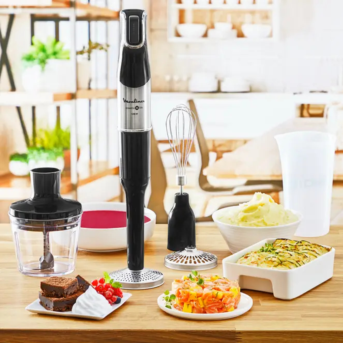 Un frullatore a immersione e accessori, con cibo su piatti e ciotole in cucina.