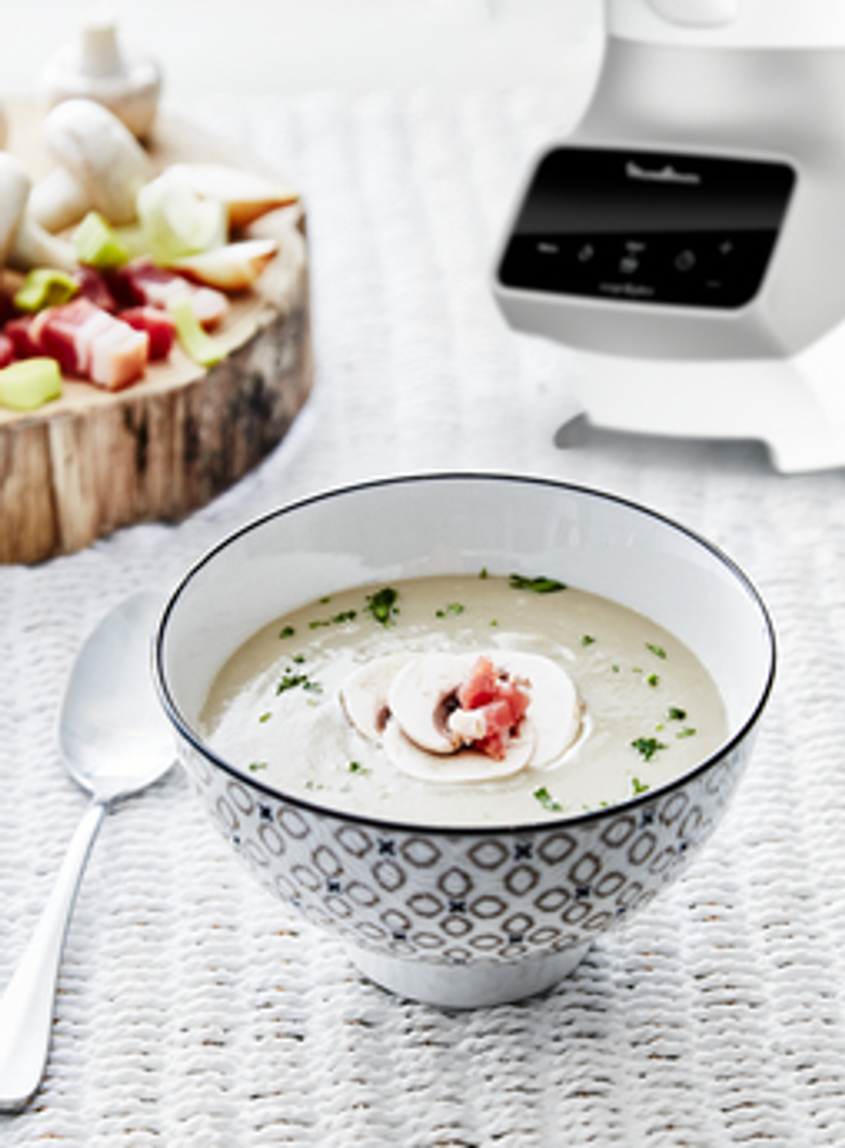 Una ciotola di zuppa con guarnizione di funghi e prosciutto, un cucchiaio e un robot da cucina.