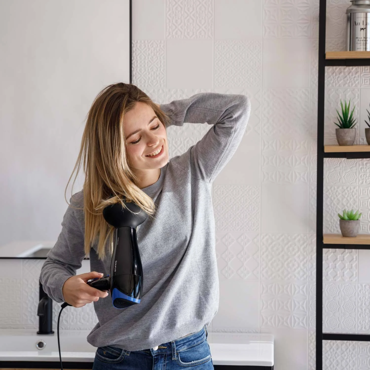 Mujer secando su cabello con un secador, sonriendo en un baño blanco.