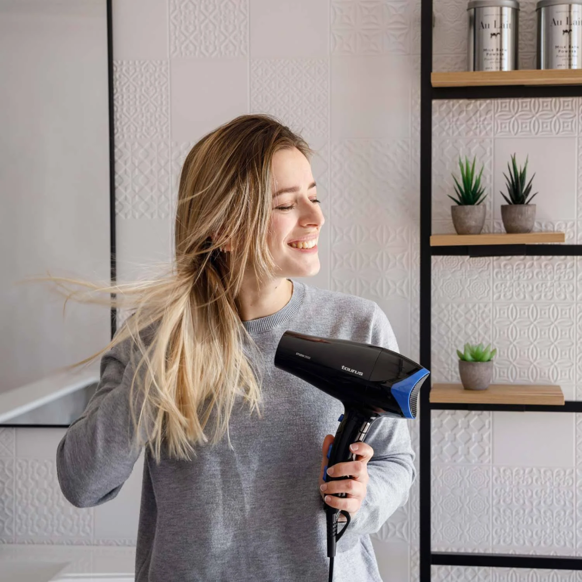 Mujer sonriendo, secándose el pelo con un secador de pelo negro y azul. Pared con azulejos blancos al fondo.