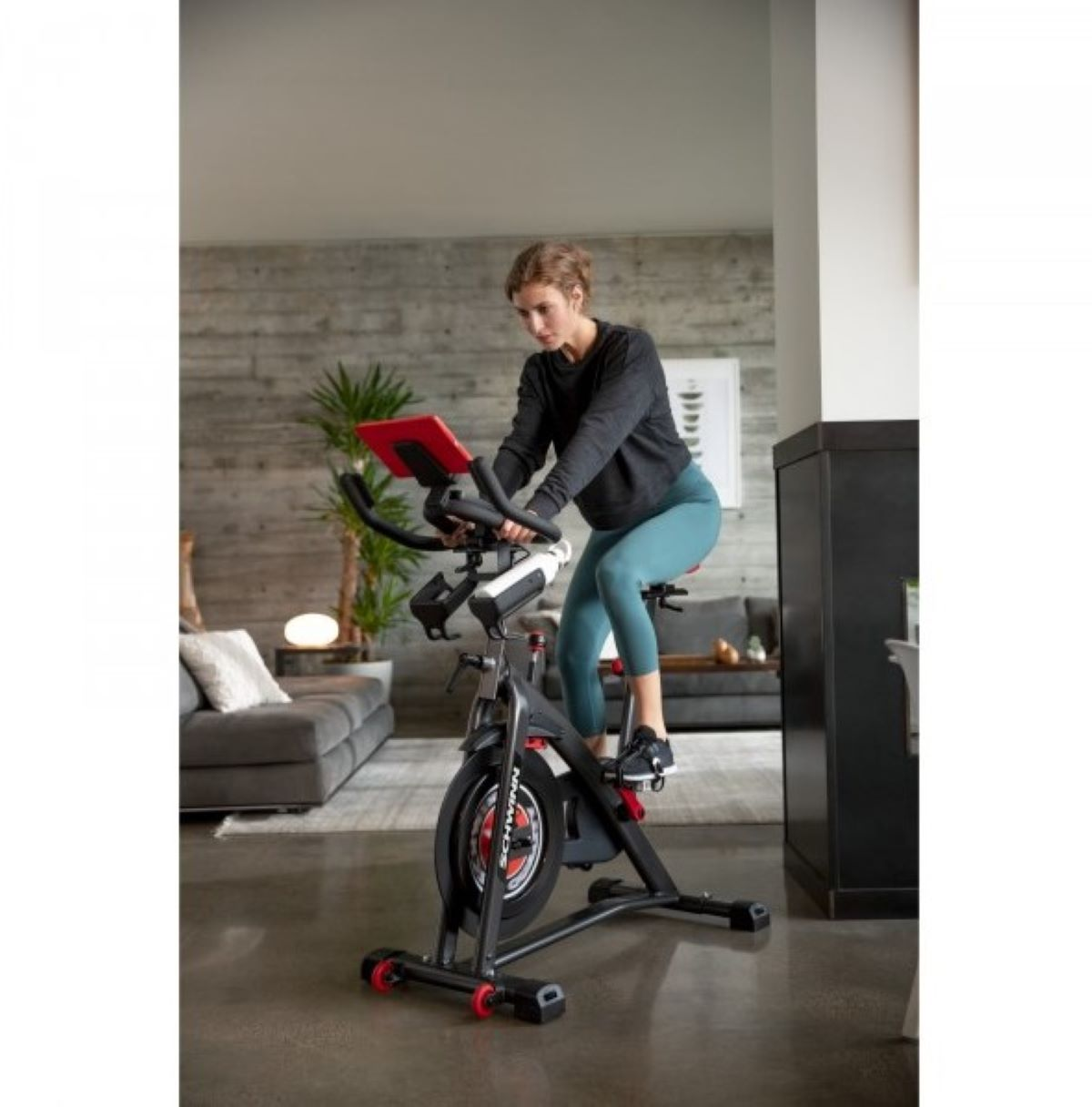 Kobieta na rowerze stacjonarnym w salonie. Rower jest czarny i czerwony.