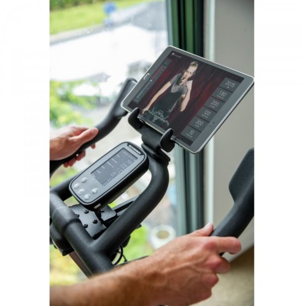 Osoba na rowerze stacjonarnym korzystająca z tabletu. Tablet wyświetla film z ćwiczeniami. Ręce trzymają rower.