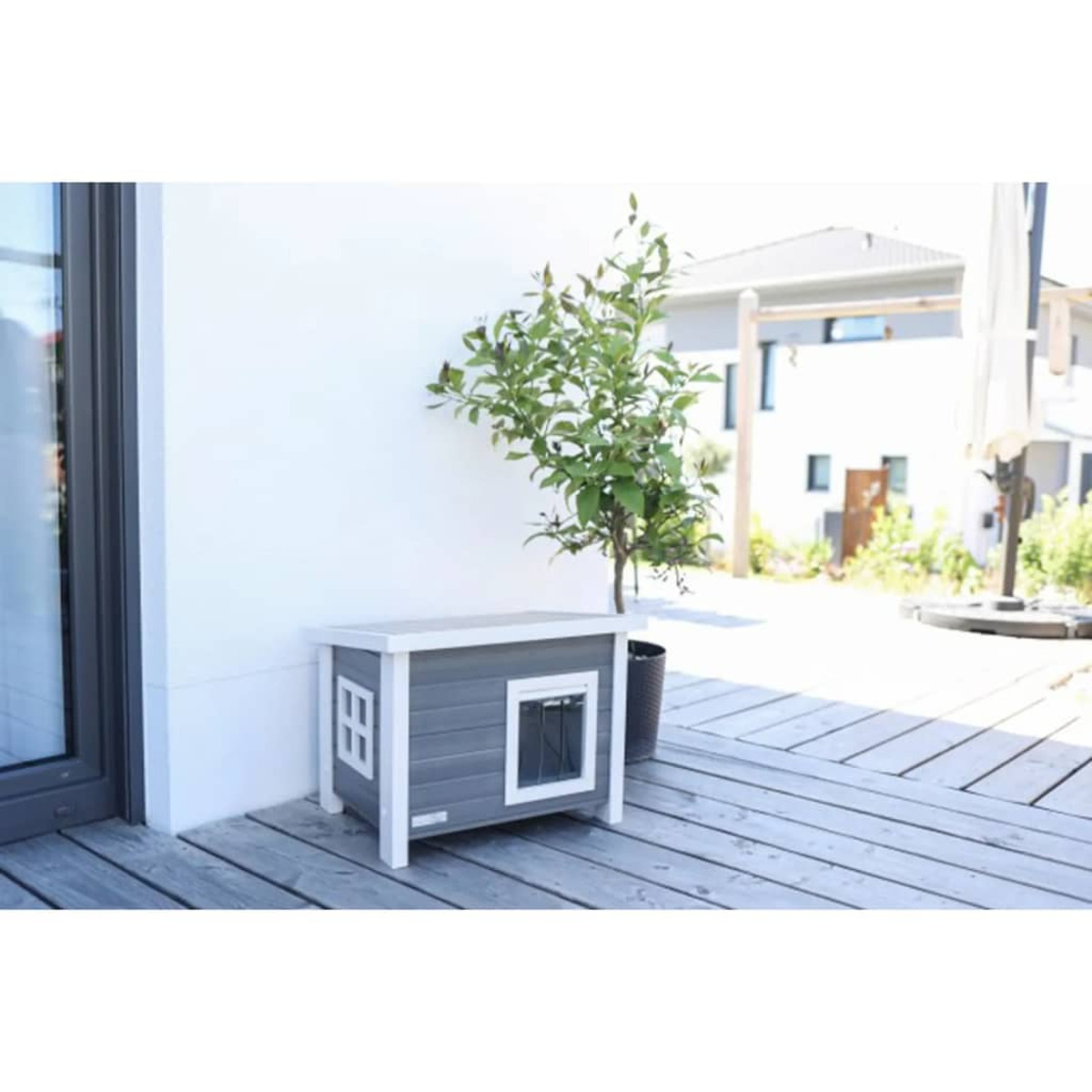 Una cuccia su una terrazza di legno, vicino a un muro bianco e un albero.
