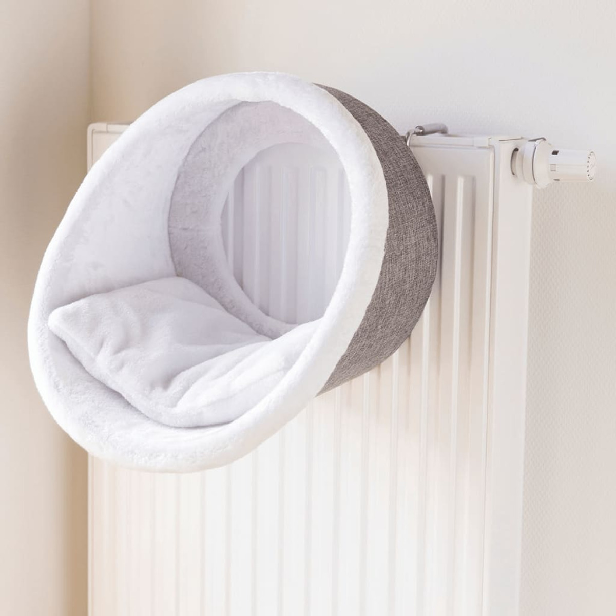 Cama para gatos montada en un radiador. Es gris y blanco con una almohada dentro. La cama es redonda.