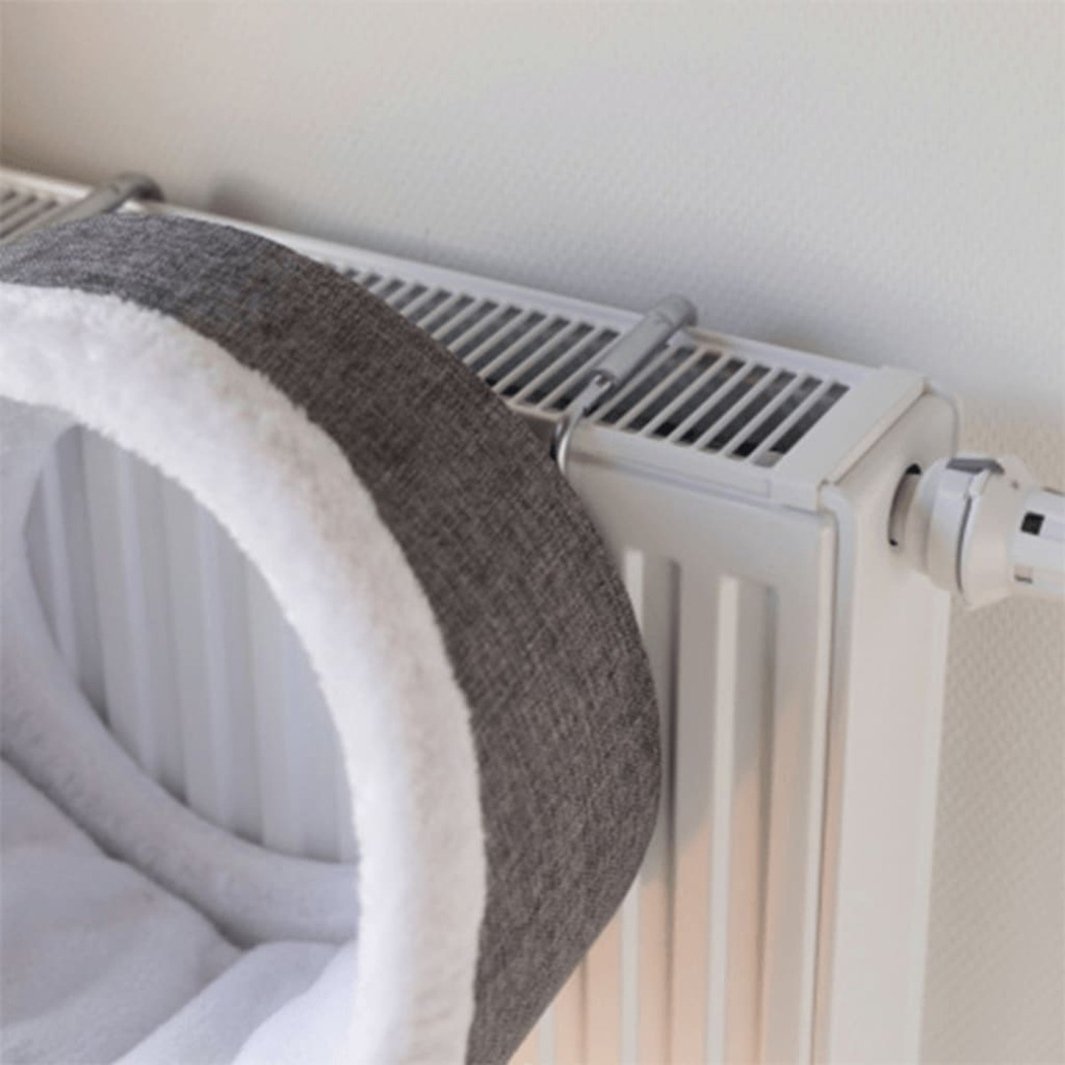 Una cama para gatos sujeta a un radiador blanco. La cama es gris con ribete blanco.