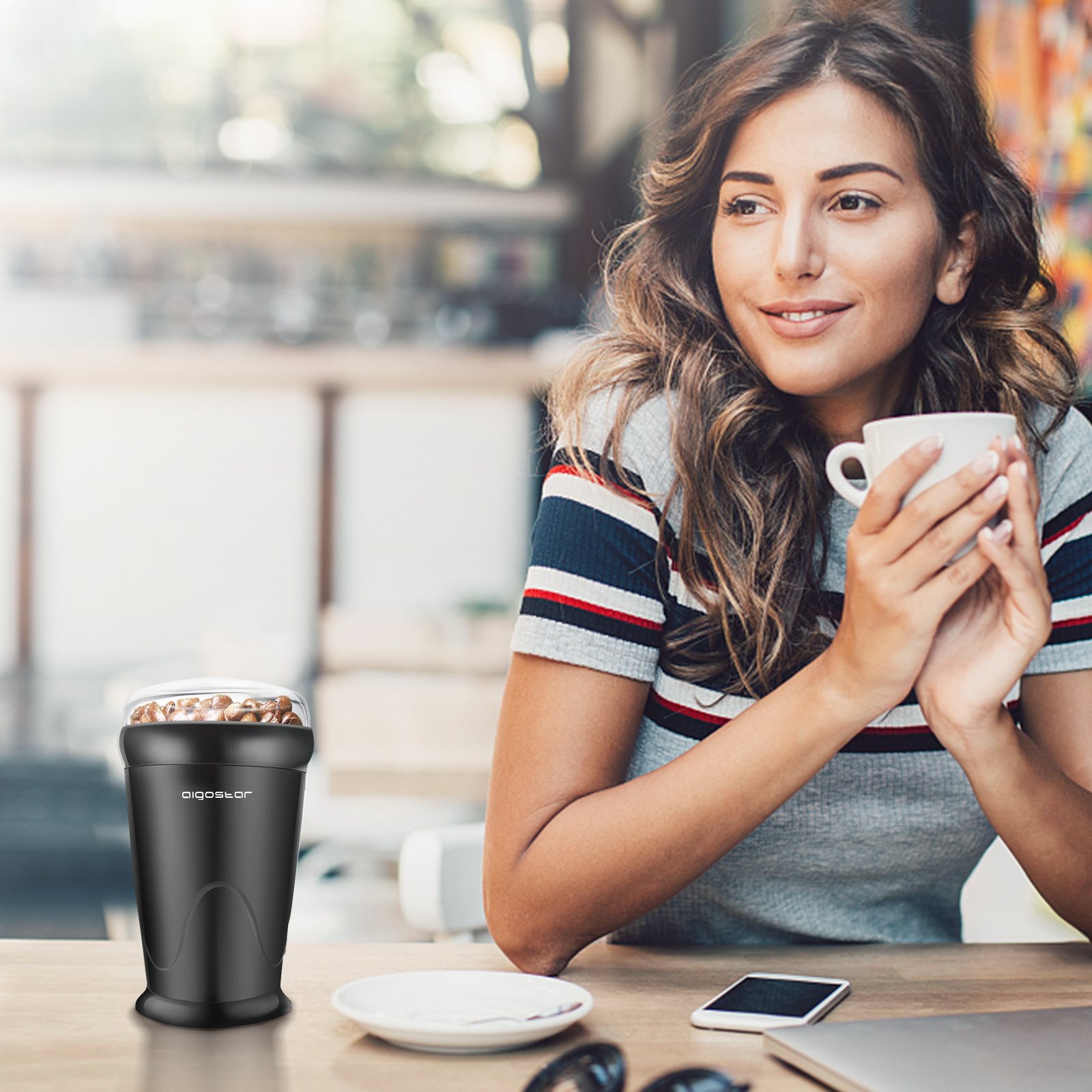 Frau hält eine Tasse, neben einer Kaffeemühle, mit Telefon und Sonnenbrille auf dem Tisch.