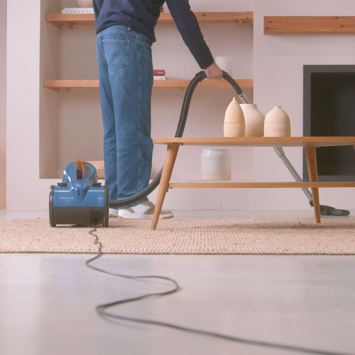 Osoba odkurzająca dywan niebieskim odkurzaczem, widoczna rura odkurzacza.