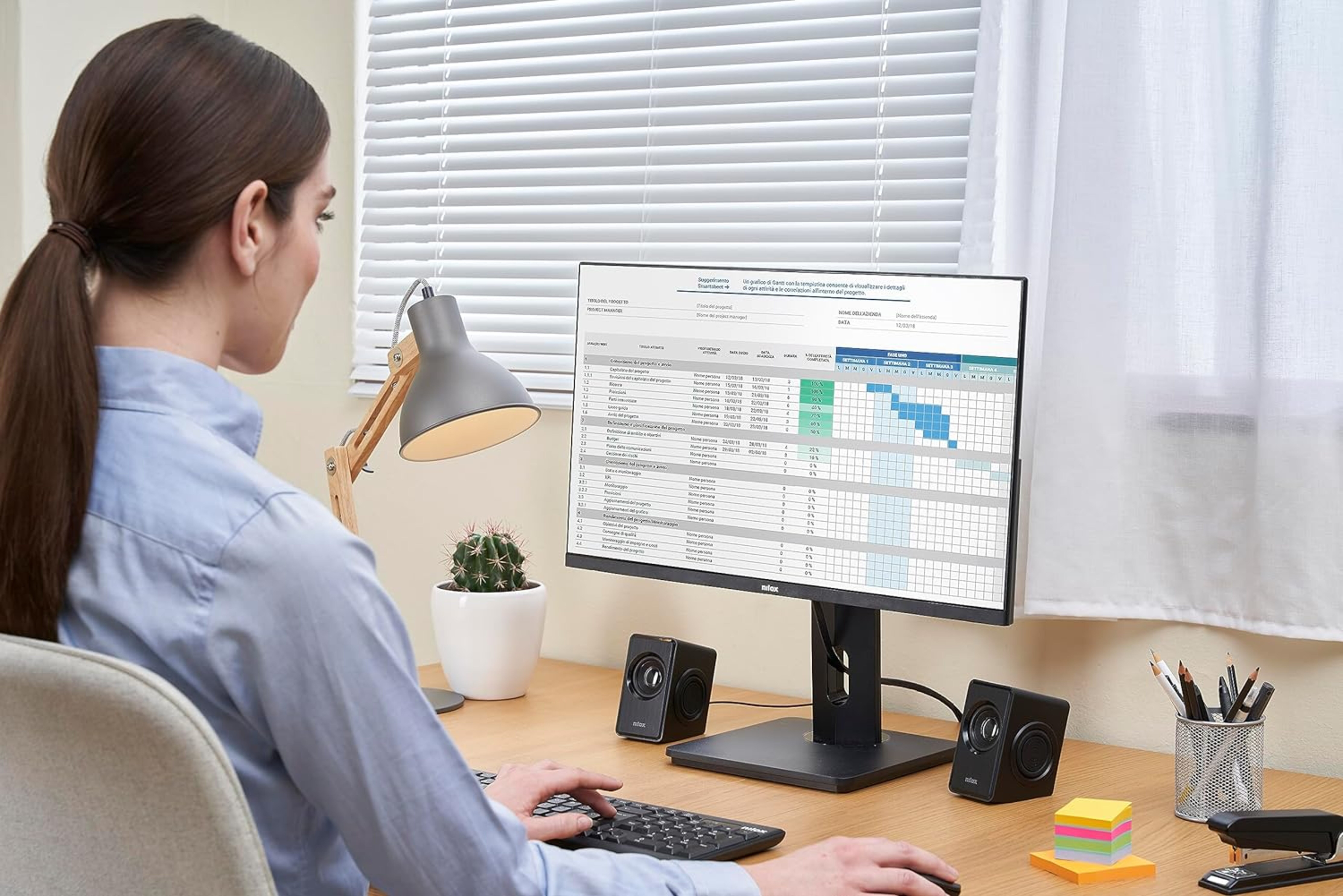Mujer trabajando en escritorio con computadora, monitor muestra hoja de cálculo, lámpara y cactus.