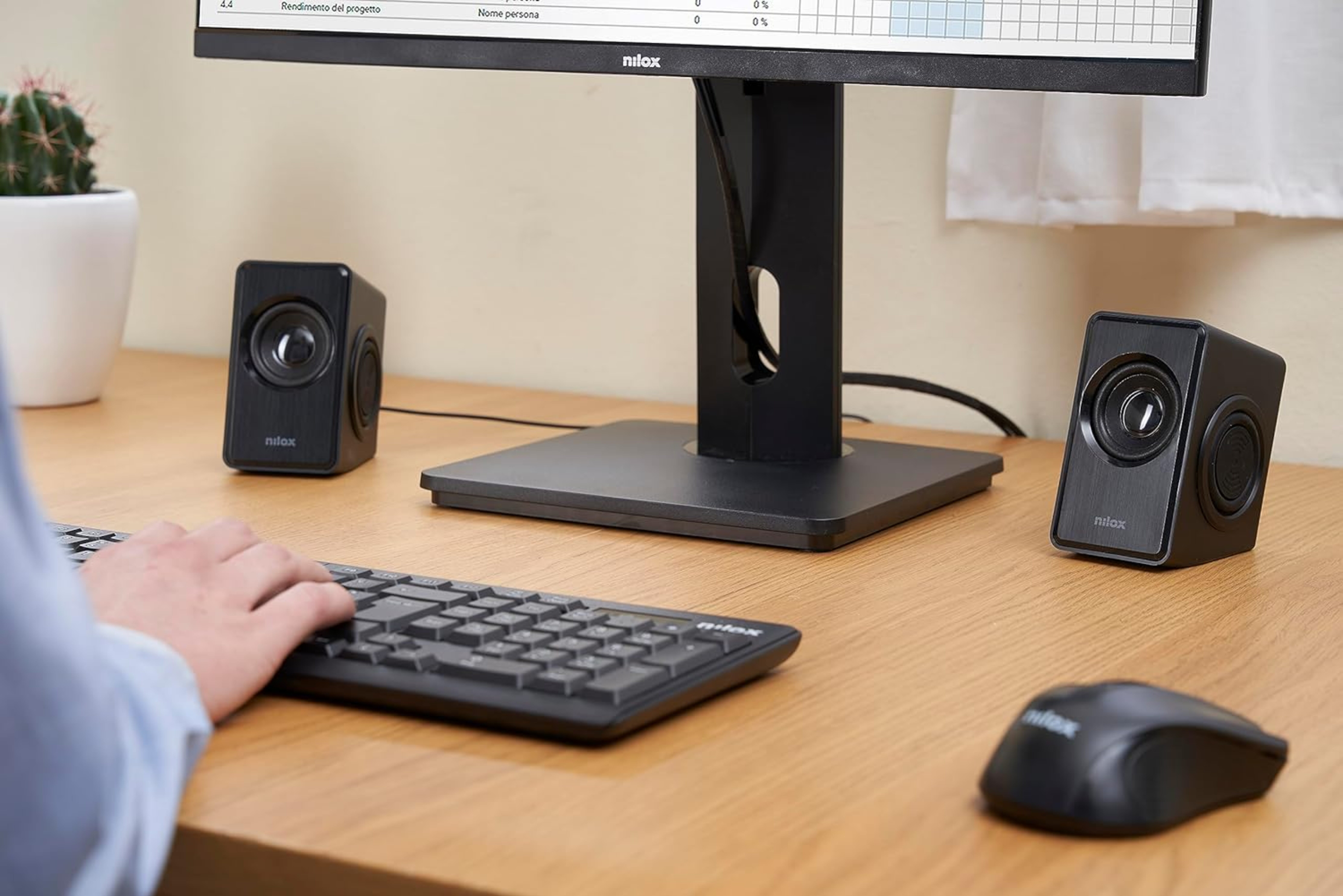 Dos altavoces de ordenador, teclado y ratón sobre un escritorio. Un monitor muestra una hoja de cálculo en el fondo.