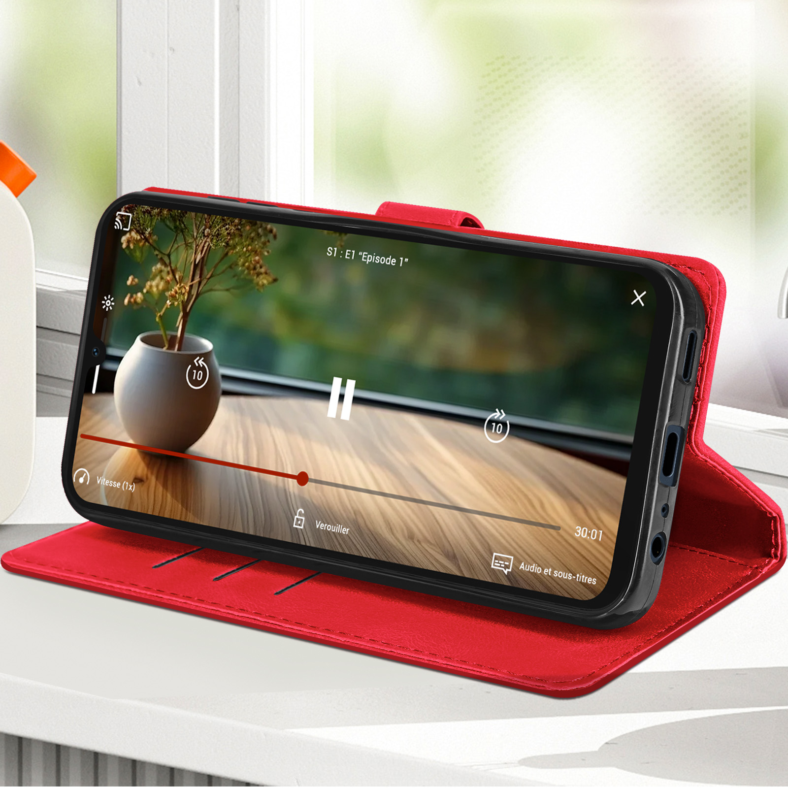 Telefono con film, in custodia rossa, su tavolo legno con pianta in vaso.