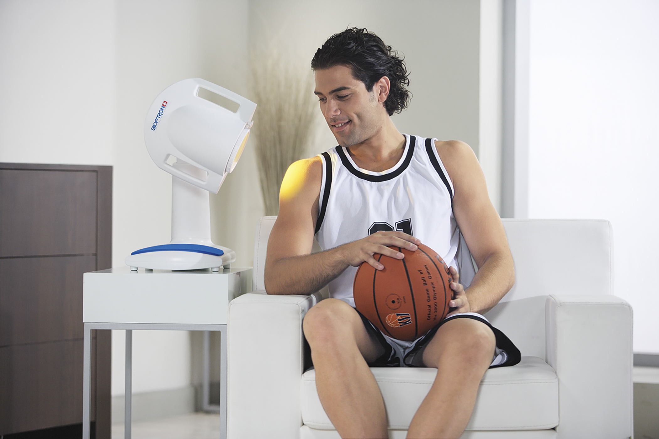 Ein Mann sitzt und hält einen Basketball in einem weißen Raum in der Nähe eines Lichttherapiegeräts.