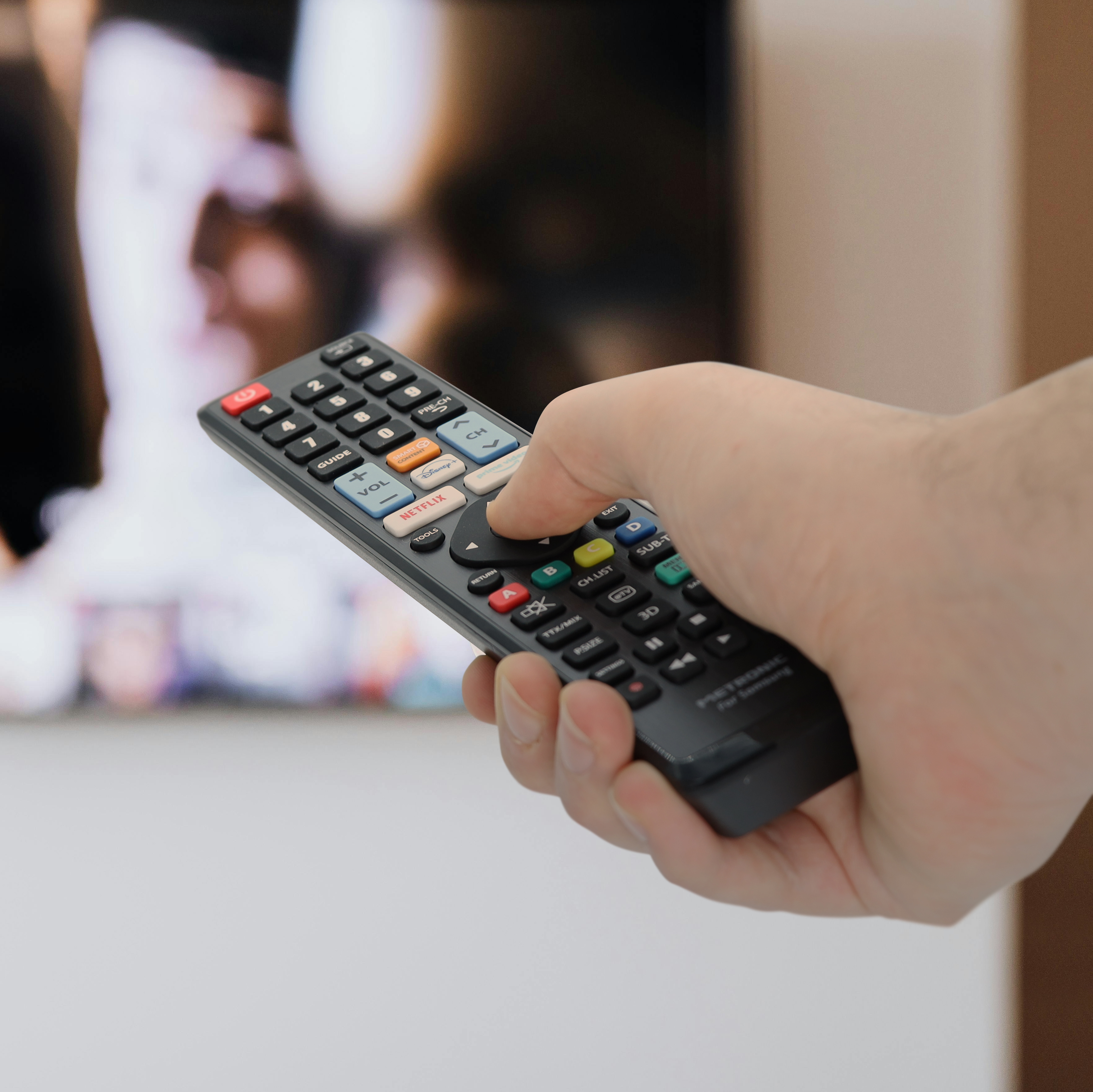 Una mano sosteniendo un control remoto de TV negro. El control remoto apunta a una pantalla de televisión borrosa.