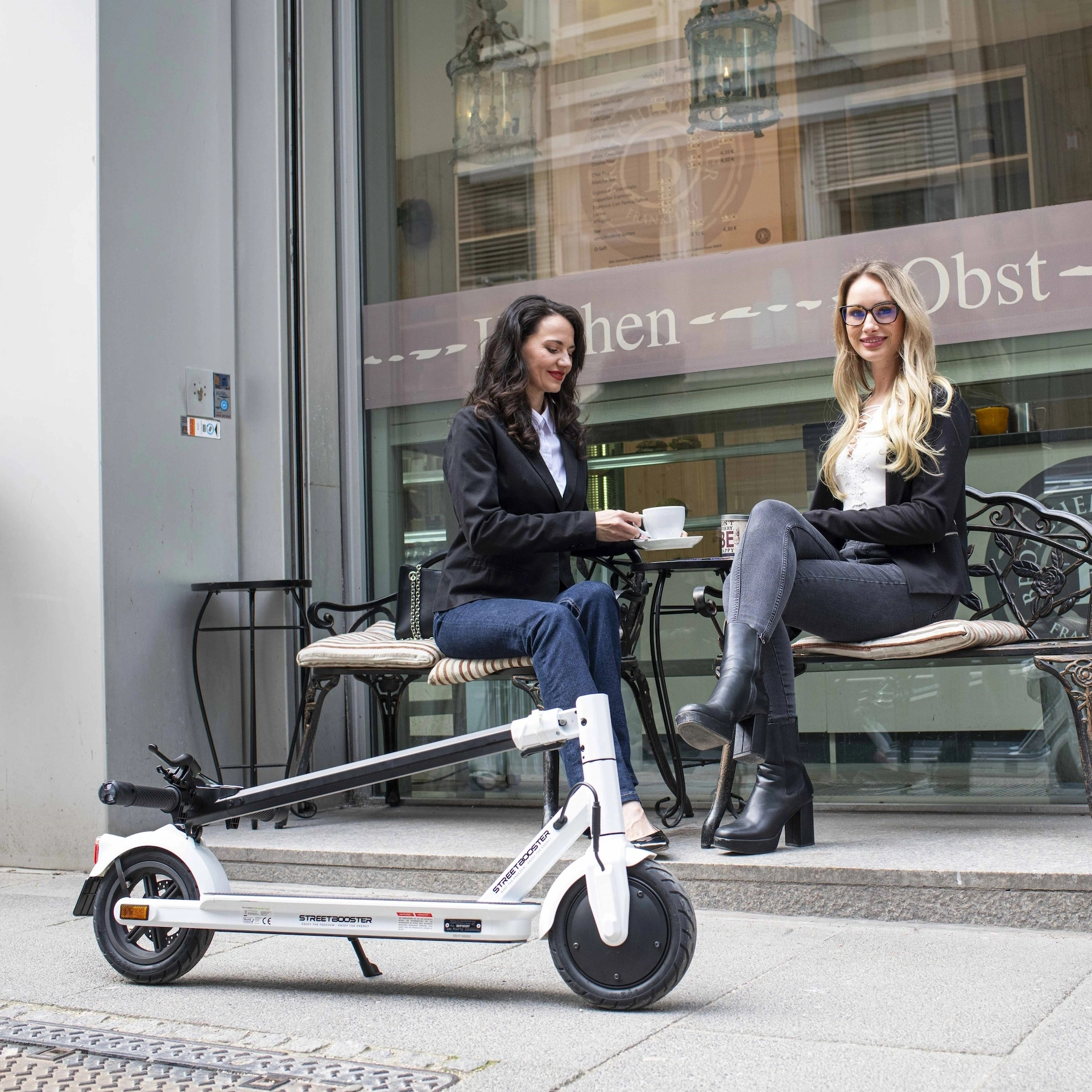 Zwei Frauen sitzen neben einem weißen Roller vor einem Geschäft.
