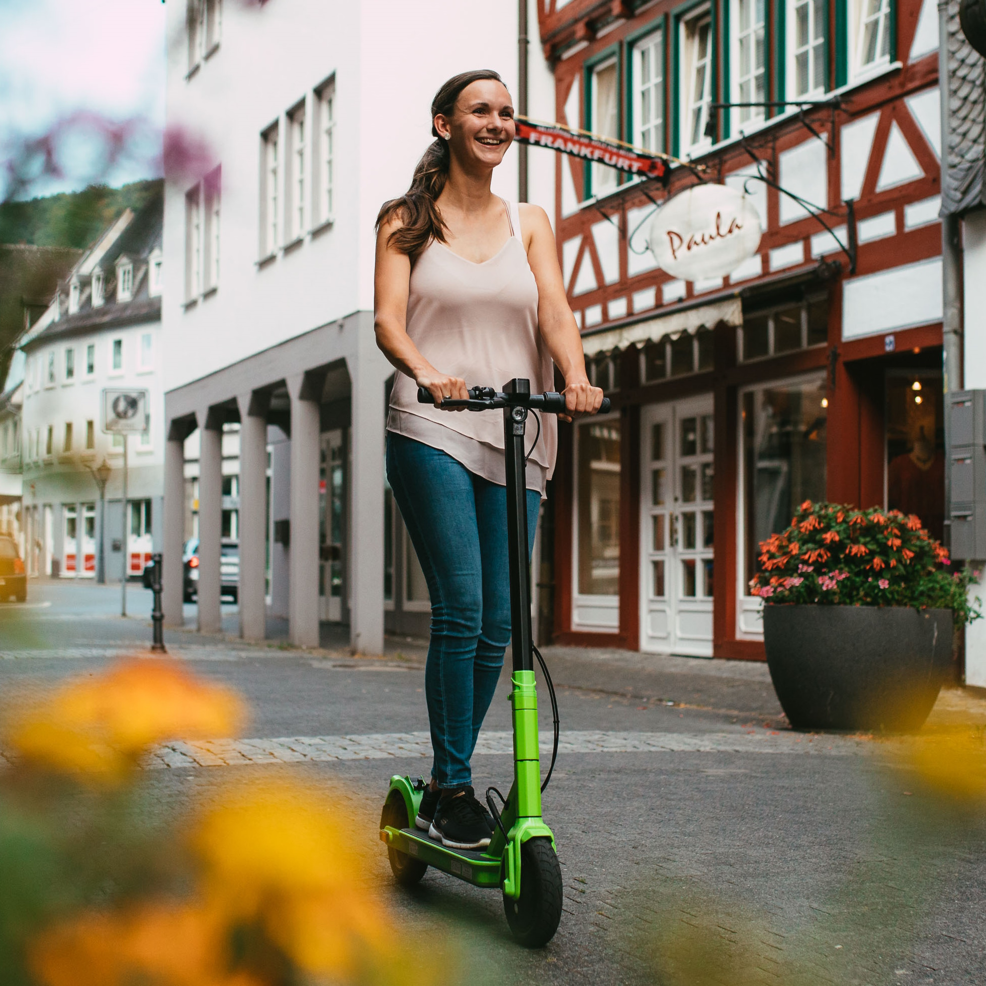 Frau fährt einen grünen Roller auf der Straße. Gebäude und Blumen sind im Hintergrund.