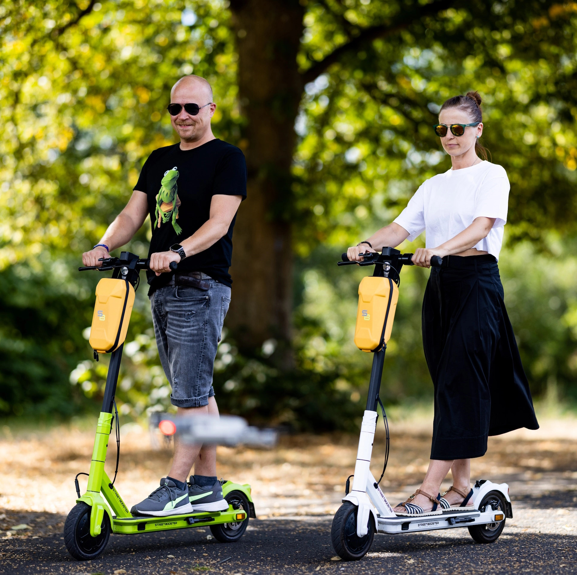 Zwei Personen auf Rollern, einer grün, einer weiß, fahren auf einem Weg. Sonnenbrillen und ein von Bäumen gesäumter Weg sind sichtbar.