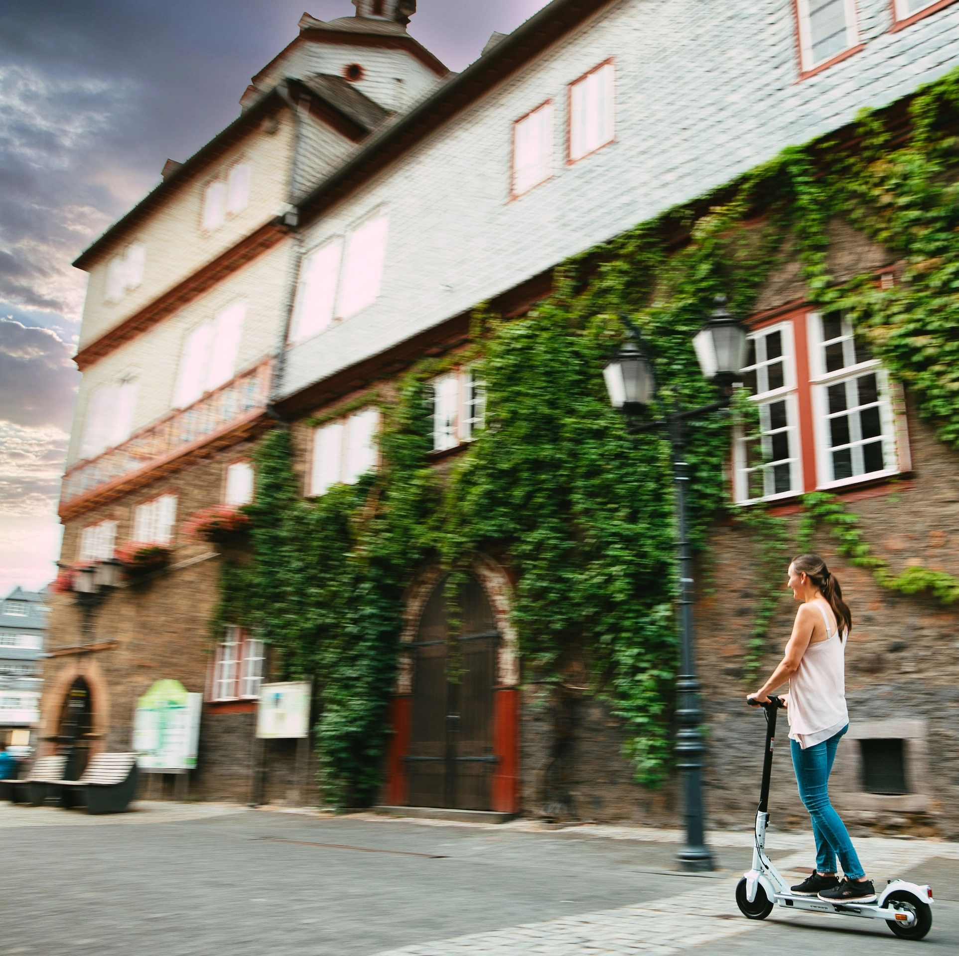 Eine Frau auf einem Roller fährt eine Straße entlang, rechts ein Gebäude und bewölkter Himmel.