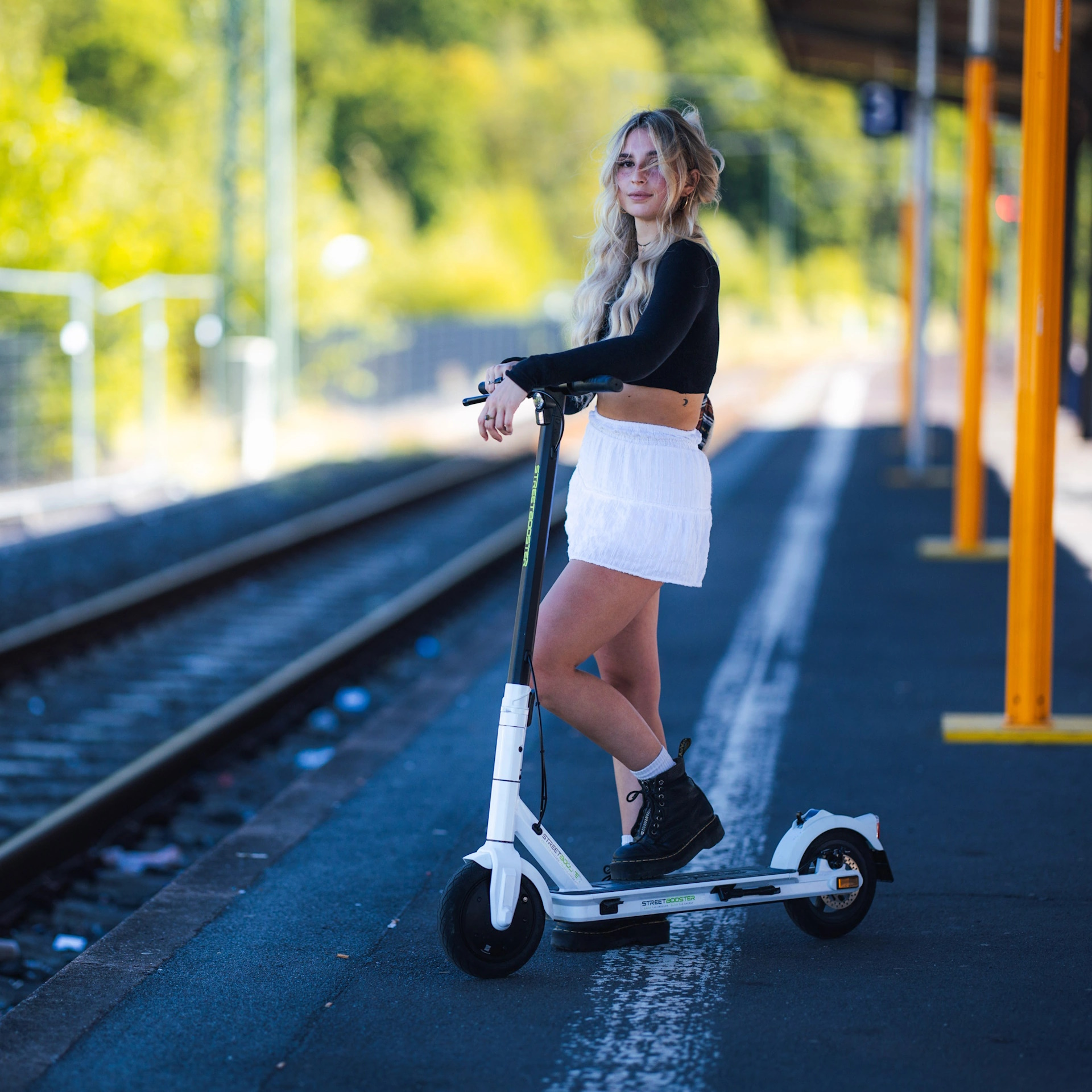 Blonde Frau auf einem weißen Roller am Bahnhof. Sie trägt ein schwarzes Oberteil und einen weißen Rock.